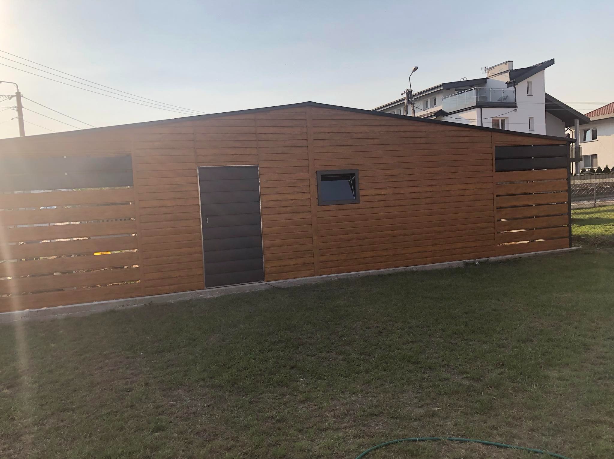 Nowoczesny, drewnopodobny garaż z płaskim dachem i ciemnymi akcentami, w tym bocznymi panelami i drzwiami, na tle zielonego trawnika i zabudowań mieszkalnych.