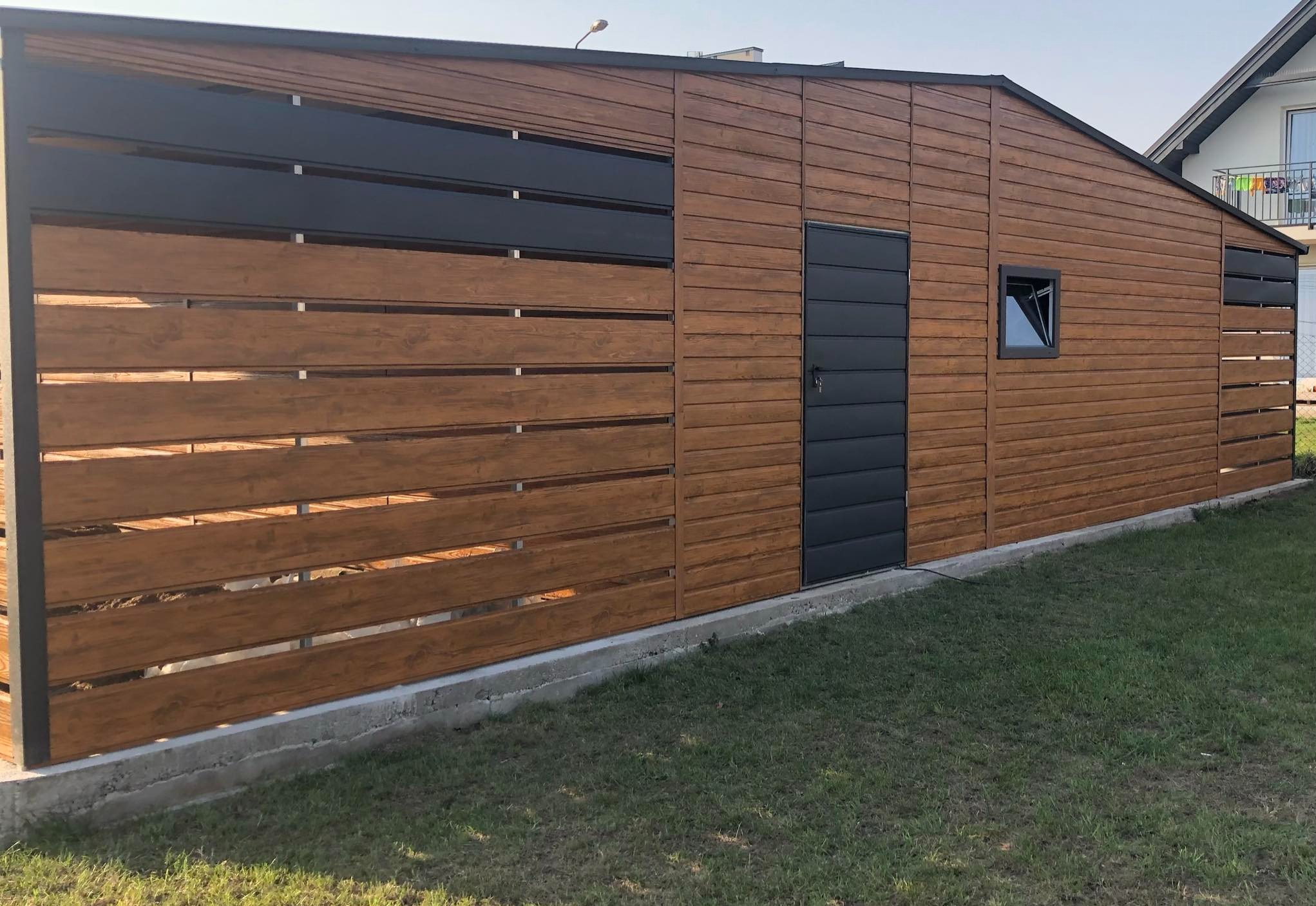 Nowoczesny garaż z drewnianą elewacją i metalowymi akcentami, zintegrowany z ogrodzeniem o poziomym układzie desek, osadzony na betonowej podmurówce na tle zielonego trawnika.