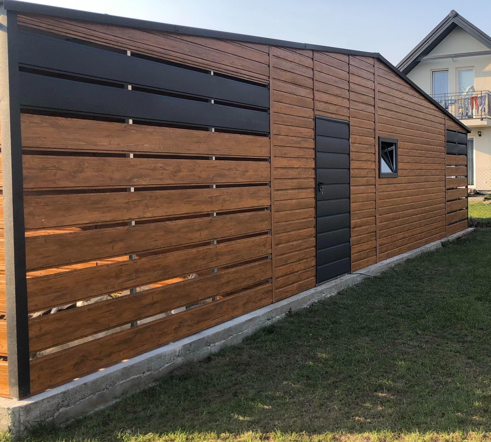 Nowoczesny, drewniany budynek gospodarczy z czarnymi akcentami, w tym drzwi i elementy ażurowe, na tle zielonego trawnika i domu jednorodzinnego.