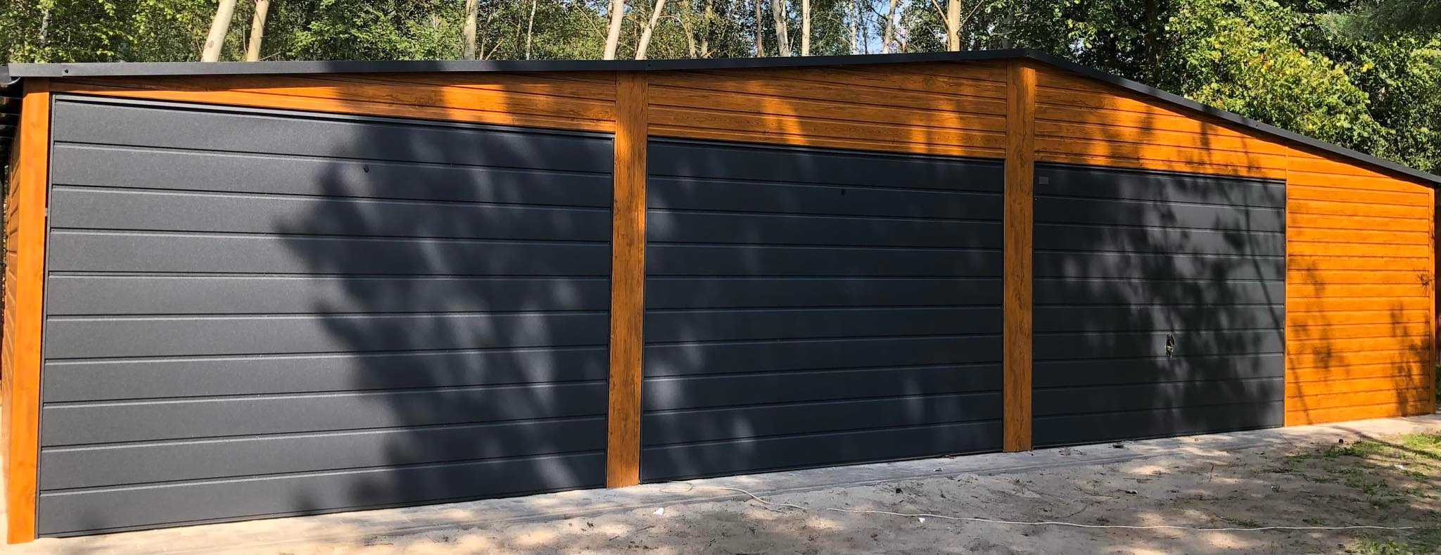 Nowoczesny, trzysegmentowy garaż z blachy w kolorze grafitowym, z drewnianymi elementami wykończeniowymi, zlokalizowany w Przyszowej, na tle drzew.