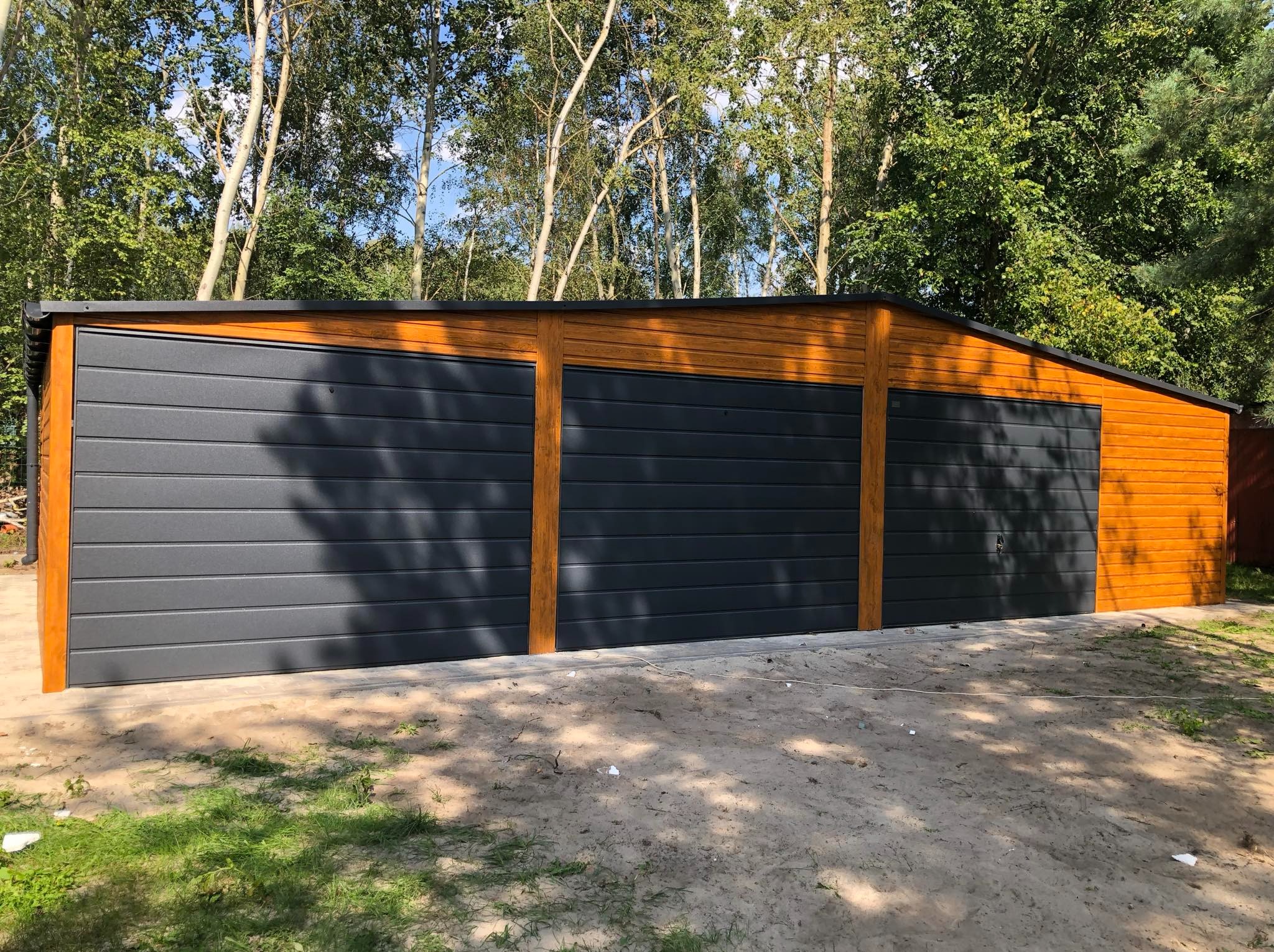 Trzysegmentowy garaż z blachy trapezowej w kolorze antracytowym, z drewnopodobnymi akcentami w kolorze miodowego dębu, na piaszczystym podłożu z widoczną trawą, otoczony drzewami.