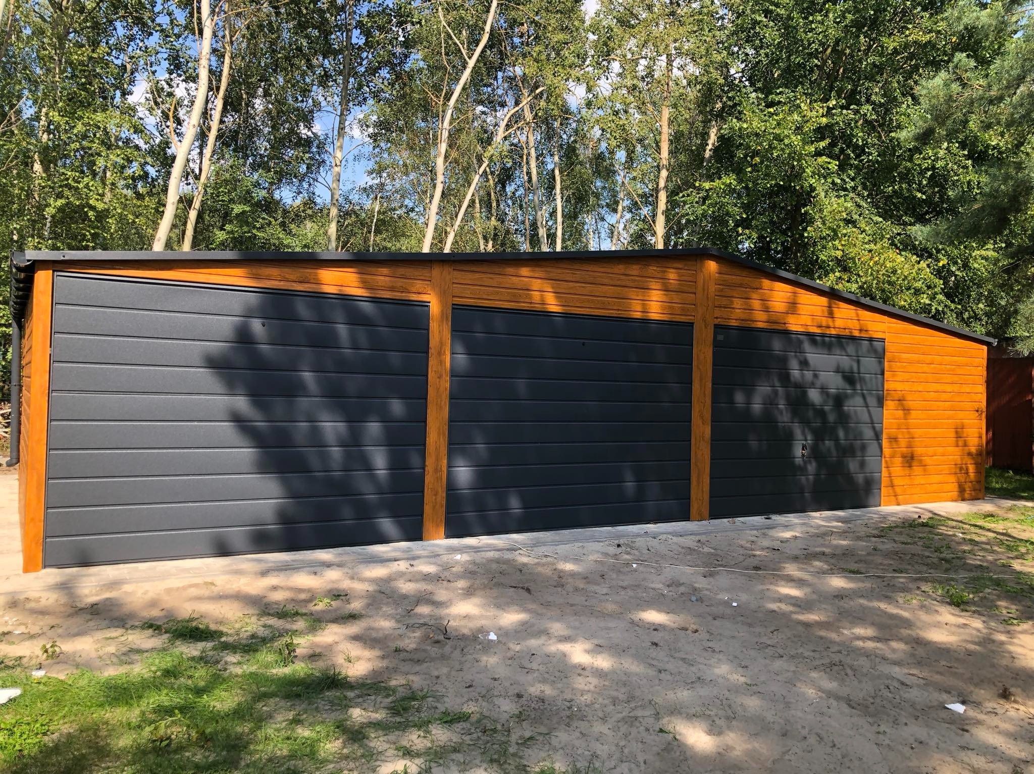 Potrójny garaż z blachy trapezowej w kolorze grafitowym, z drewnopodobnymi elementami wykończeniowymi, na piaszczystym podłożu, otoczony zielenią drzew.