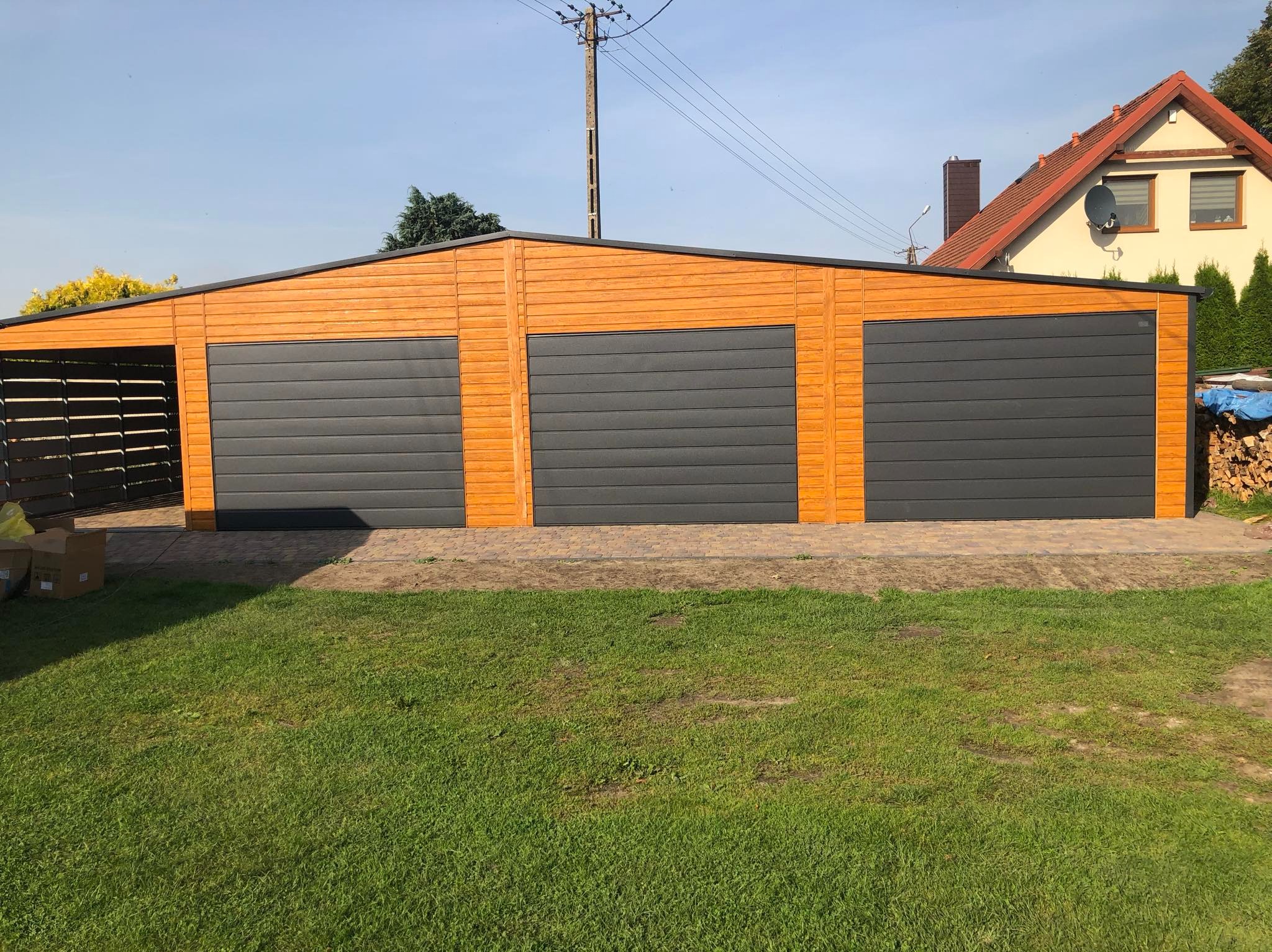 Trzysegmentowy garaż z drewnopodobną elewacją i szarymi bramami segmentowymi, widok z przodu na tle zielonego trawnika i domu jednorodzinnego.
