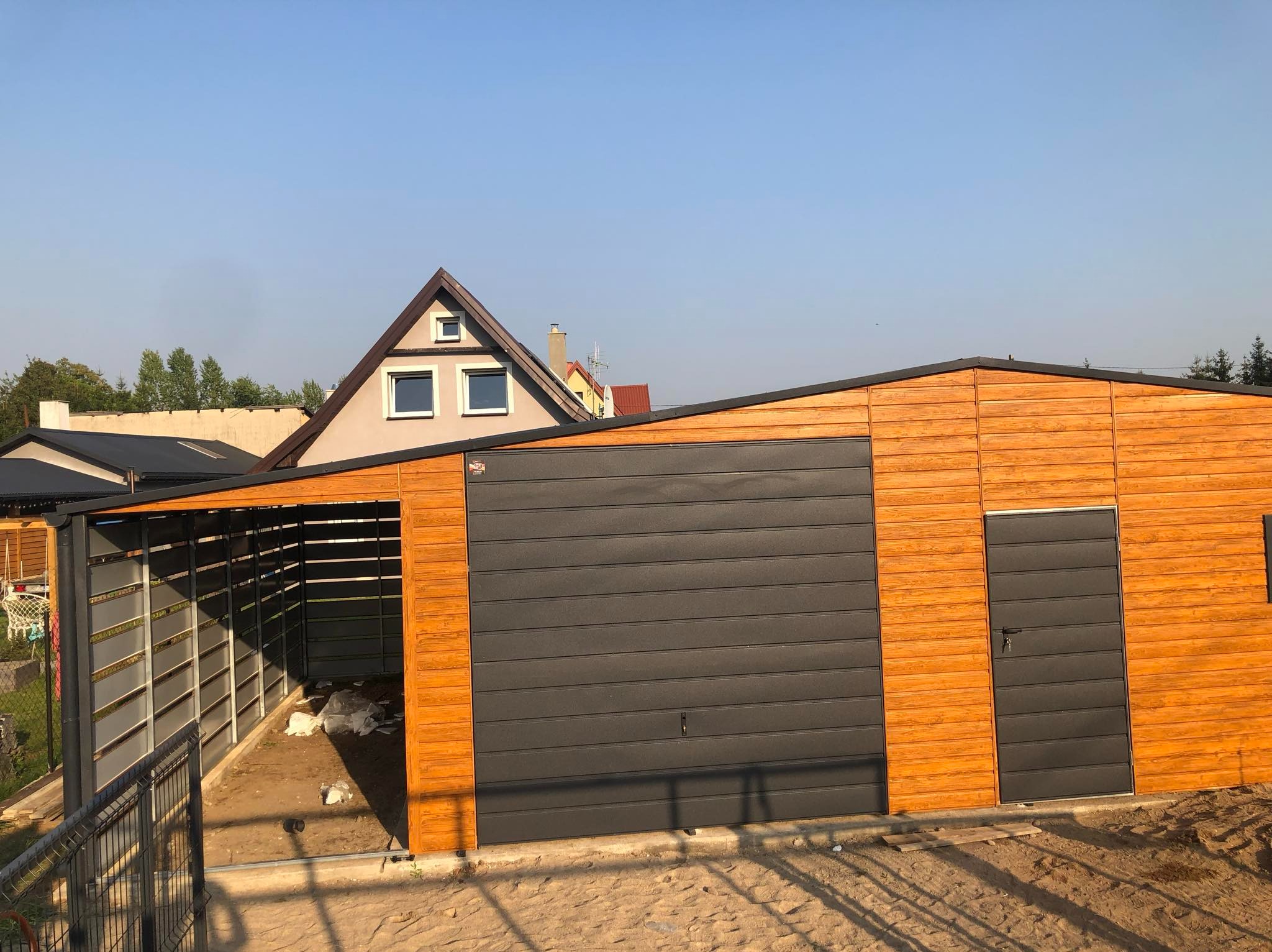 Nowoczesny garaż z blachy drewnopodobnej z szarą bramą segmentową i zadaszeniem, widok z zewnątrz na tle błękitnego nieba i zabudowań.