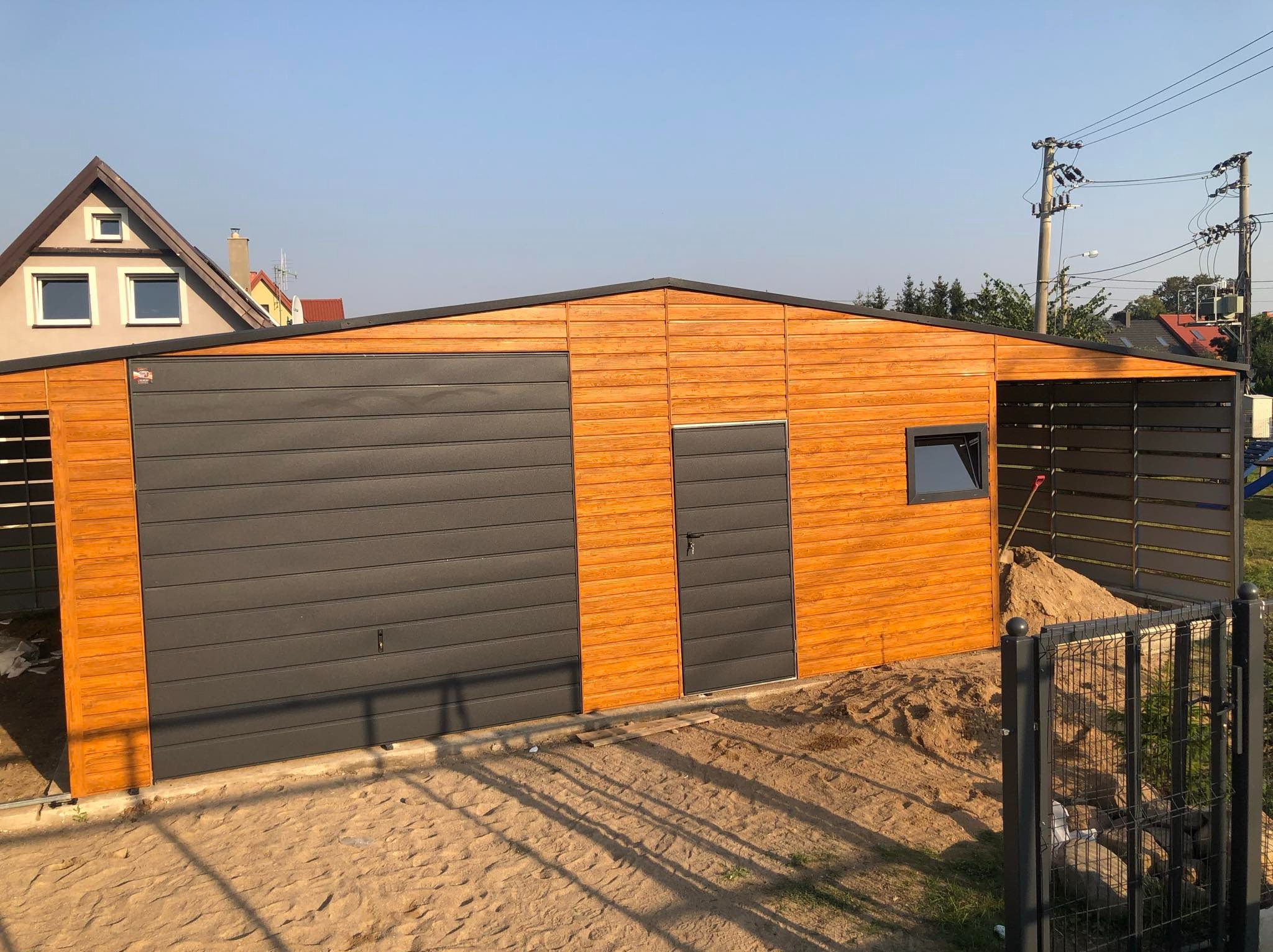 Nowoczesny garaż z blachy drewnopodobnej z szarą bramą segmentową i drzwiami wejściowymi, widoczny z zewnątrz na piaszczystym podłożu, z fragmentem ogrodzenia i domu w tle.