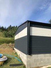 Konstrukcja z blachy trapezowej w kolorze grafitowym i kremowym, osadzona na fundamencie z bloczków betonowych, widok z boku na tle zieleni i błękitnego nieba.