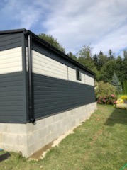 Garaż blaszany z elewacją w kolorze grafitowo-białym, osadzony na fundamencie z betonowych bloczków, widok z boku na tle zieleni.