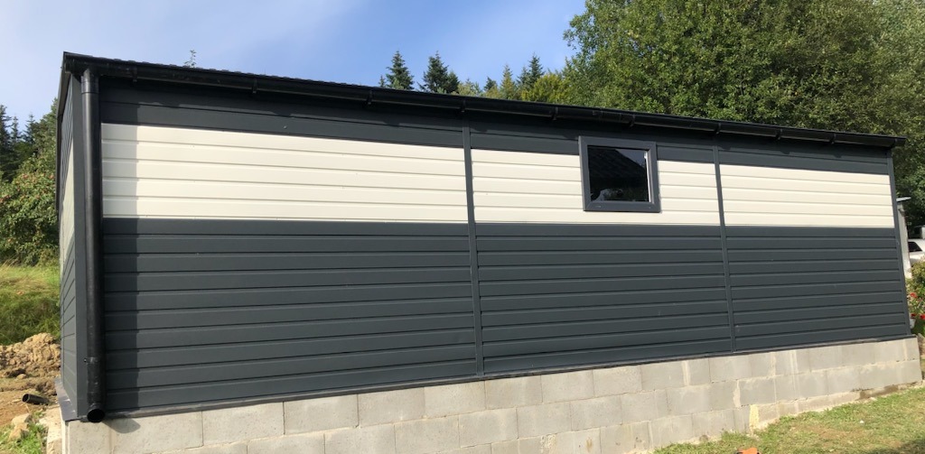 Zewnętrzny widok nowo wybudowanego, dwukolorowego budynku z blachy trapezowej z pojedynczym oknem, osadzonego na betonowej podmurówce, z widocznymi elementami orynnowania i otaczającą zielenią drzew.