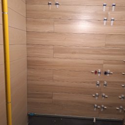 Vadym - Ściana łazienki w trakcie układania płytek imitujących drewno, z widocznymi krzyżykami dystansowymi i poziomicą przy ścianie. Widoczne wyprowadzenia instalacji wodnej.
