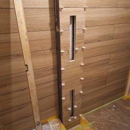 Vadym - Układanie drewnopodobnych płytek na ścianie z pionowym wnęką, widoczne krzyżyki dystansowe, poziomica oparta o ścianę, podłoga zabezpieczona folią malarską.