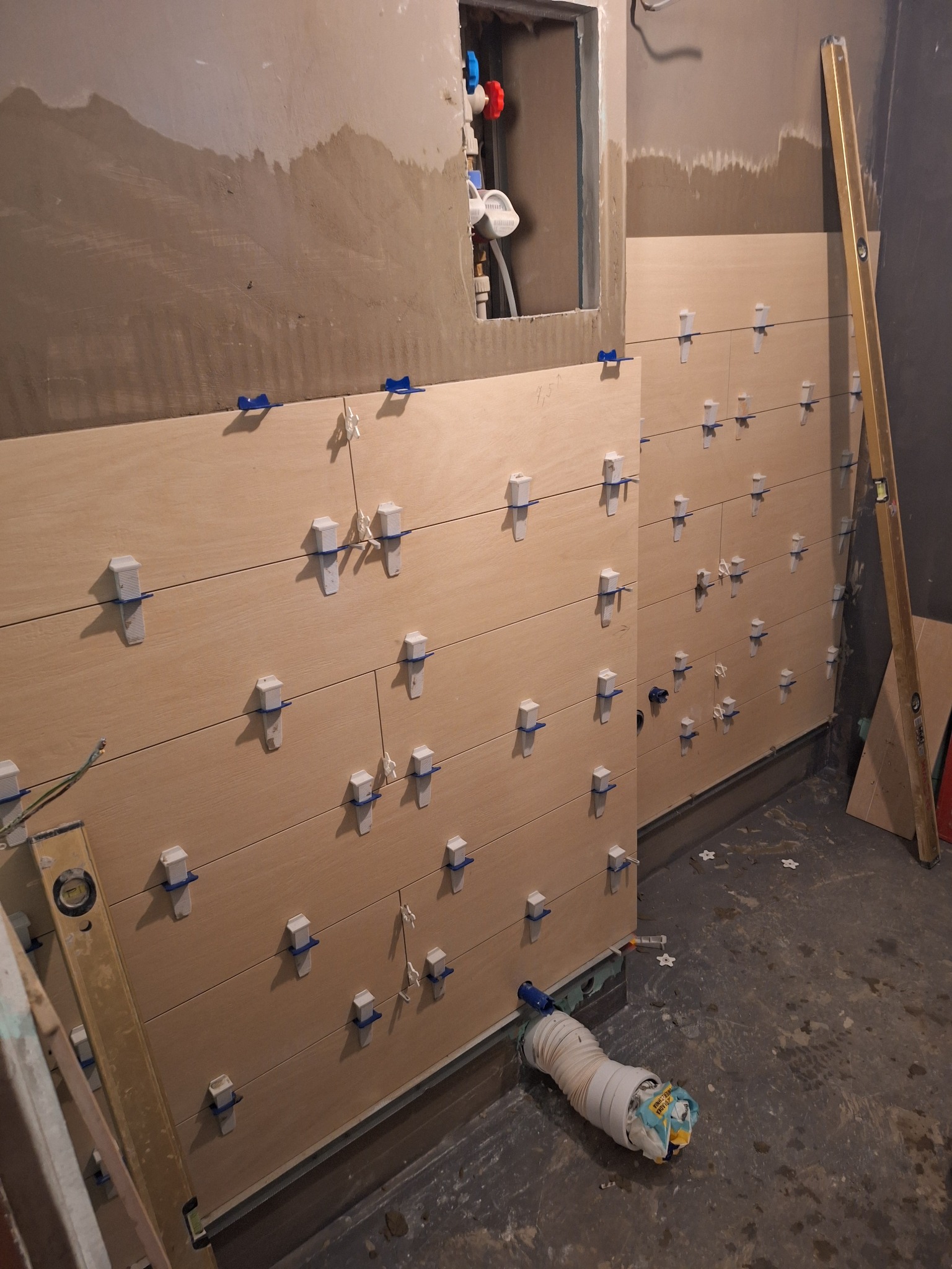 Montaż płytek imitujących drewno w łazience w Szczecinie, z użyciem poziomujących klipsów i klinów, widoczne surowe ściany i wystające rury.