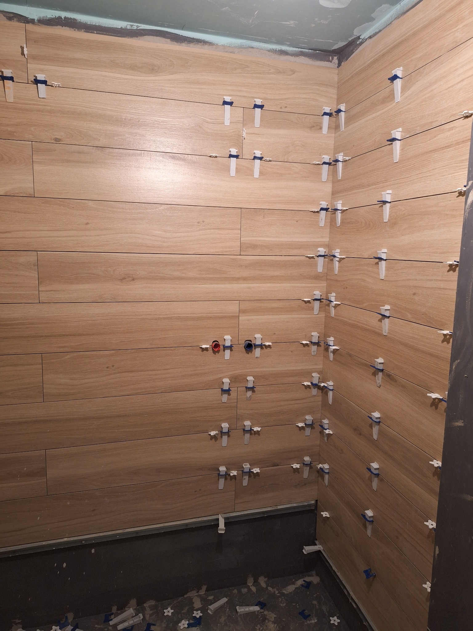 Montaż płytek imitujących drewno w łazience, widoczne systemy poziomowania płytek i przygotowane przyłącza wodne.