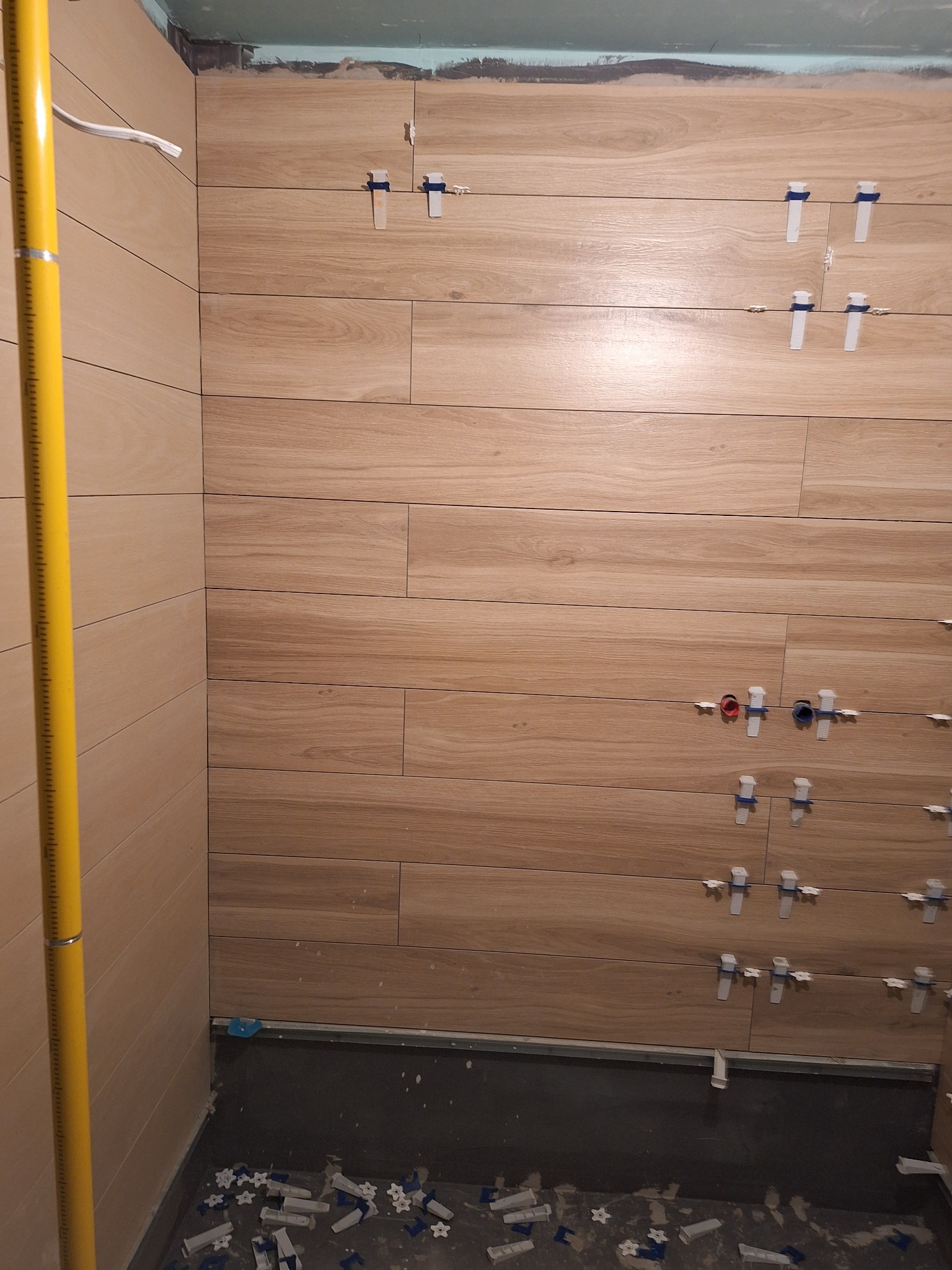 Ściana łazienki w trakcie układania płytek imitujących drewno, z widocznymi krzyżykami dystansowymi i poziomicą przy ścianie. Widoczne wyprowadzenia instalacji wodnej.