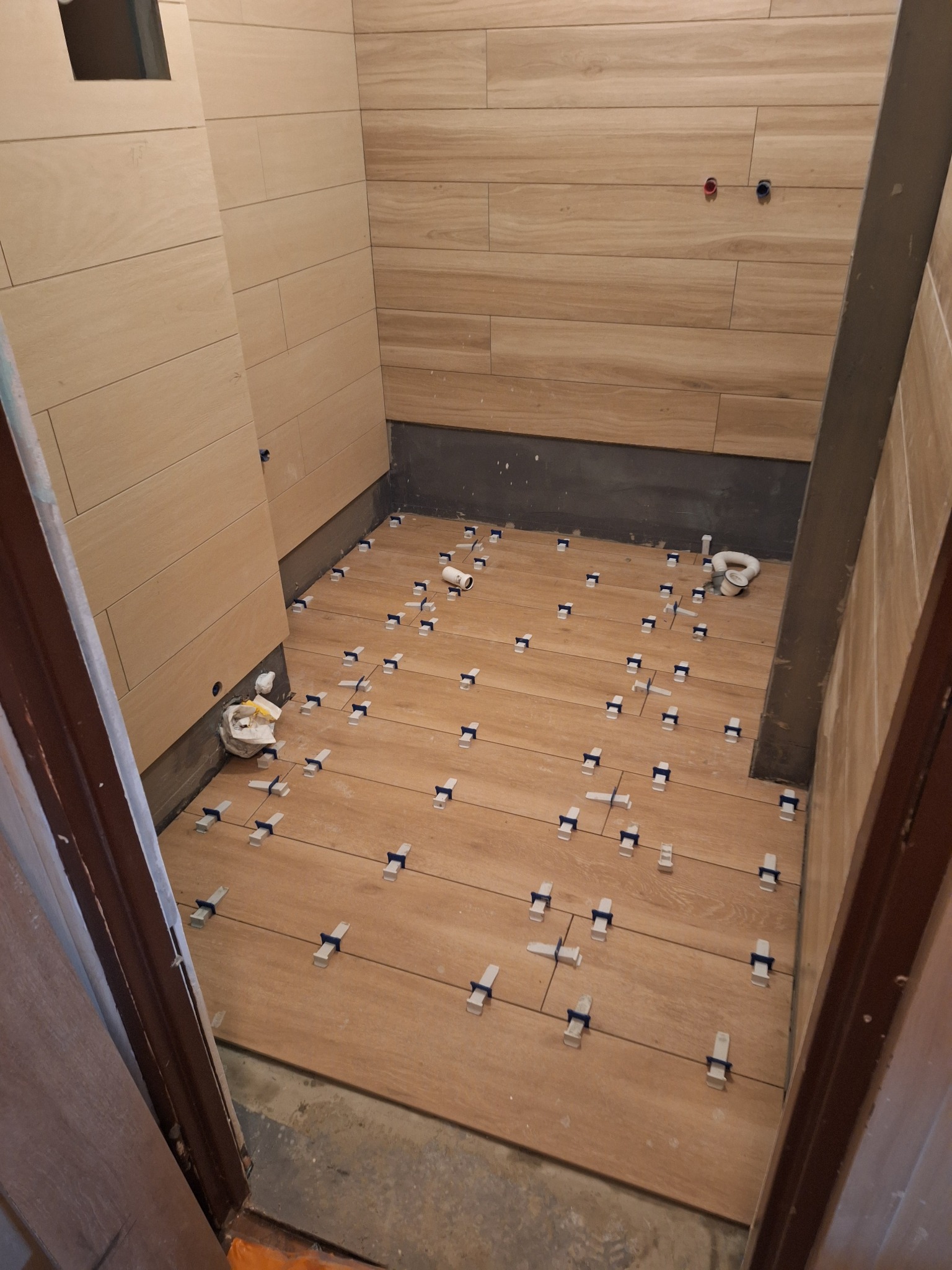 Układanie płytek podłogowych imitujących drewno w łazience, widoczne krzyżyki dystansowe i elementy kanalizacji.