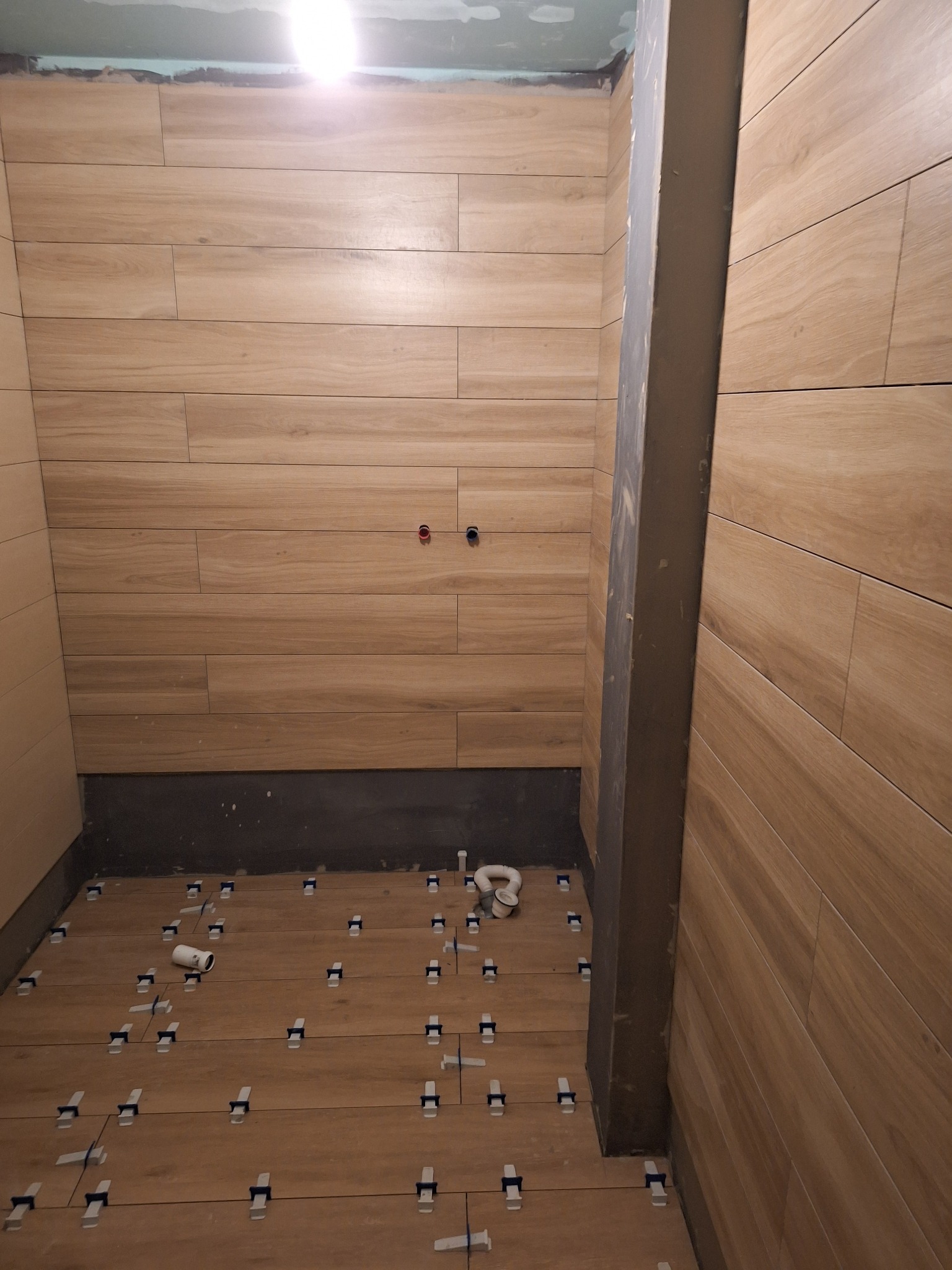 W trakcie układania płytek imitujących drewno w łazience, widoczne krzyżyki dystansowe i fragment instalacji wodnej.