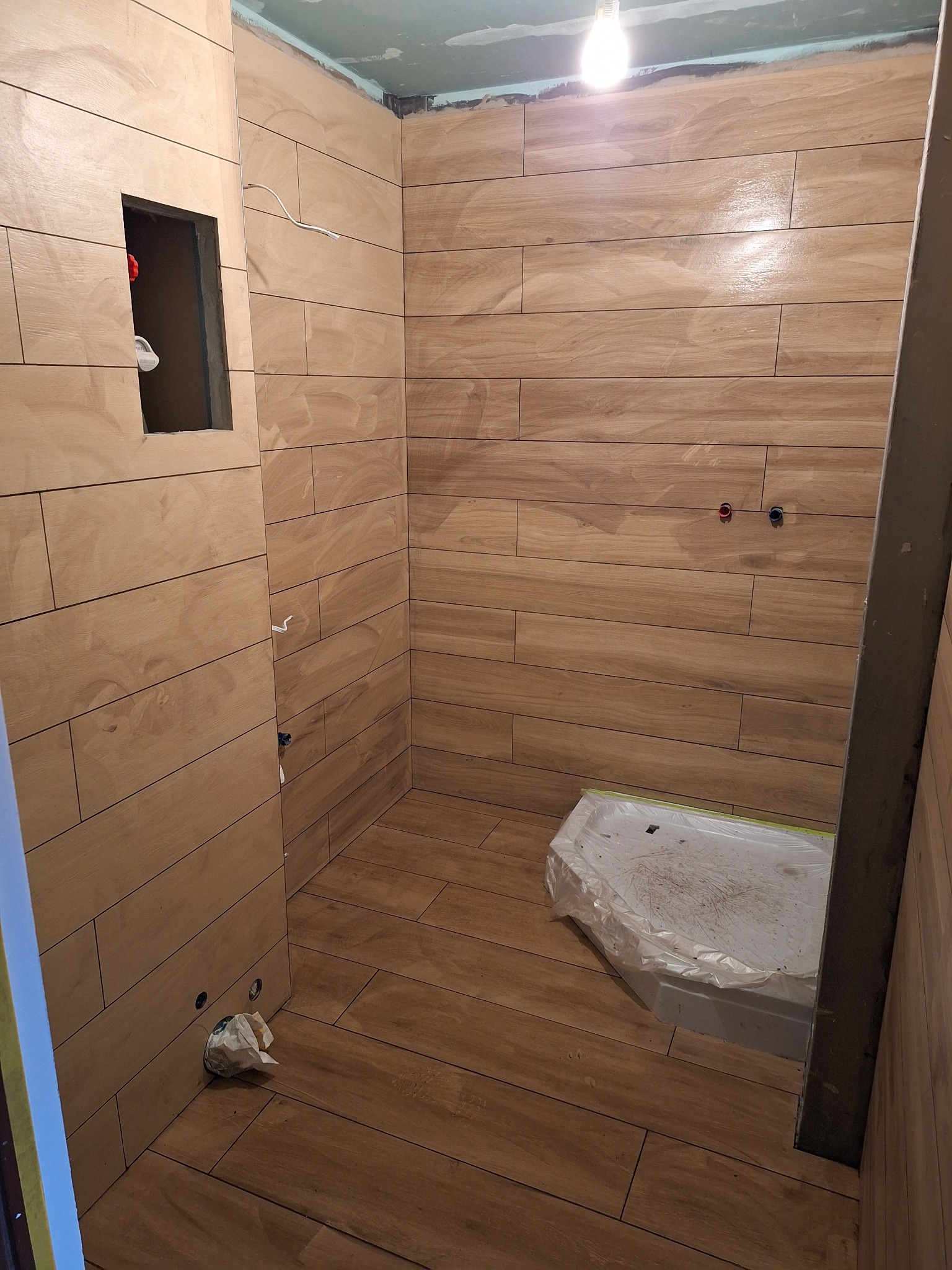 Łazienka w trakcie remontu: ściany i podłoga wyłożone brązowymi płytkami imitującymi drewno, zabezpieczony folią brodzik, widoczne punkty instalacji wodnej i elektrycznej, otwór rewizyjny.