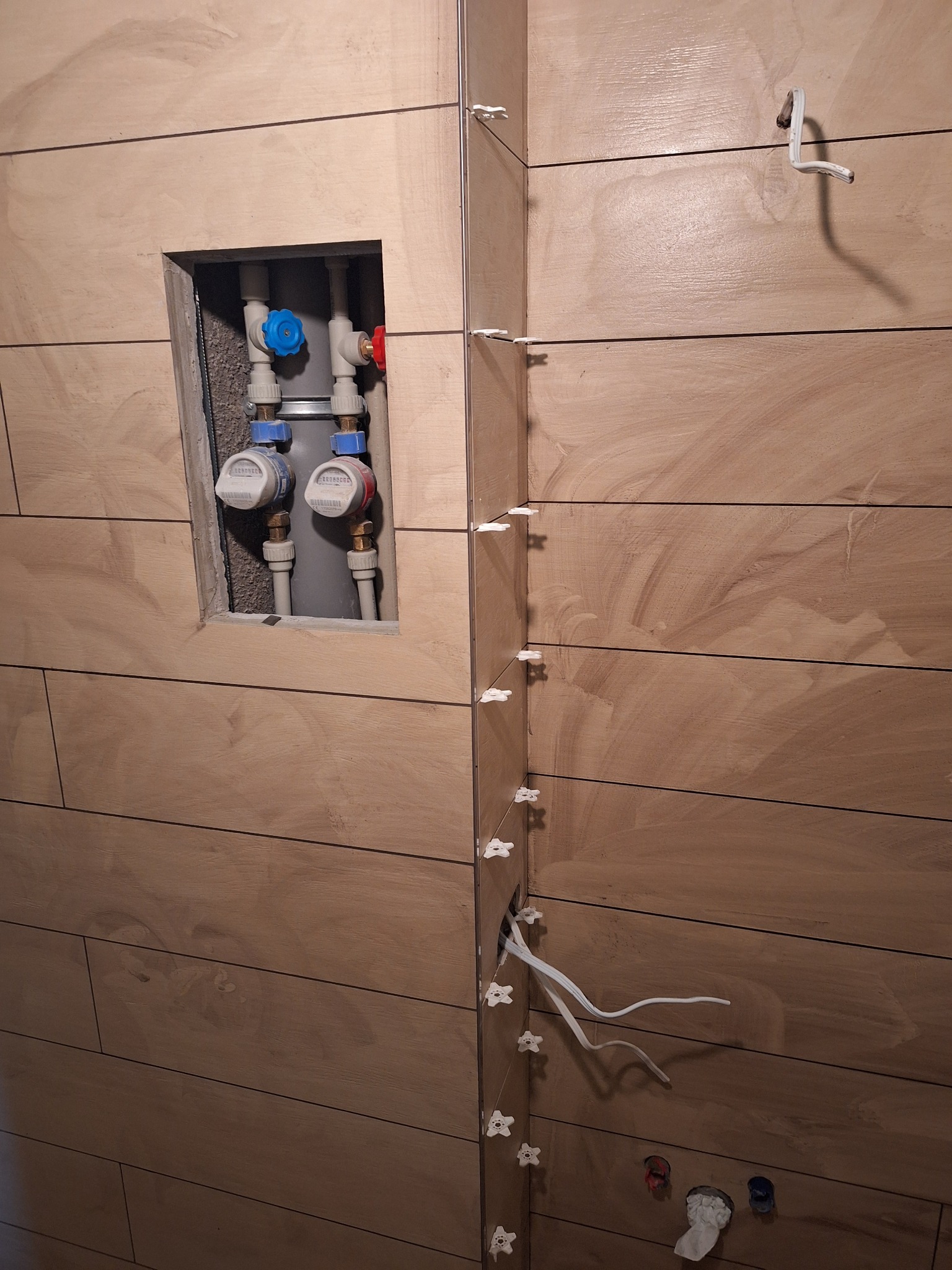 Ściana w trakcie układania płytek imitujących drewno w łazience, widoczne krzyżyki dystansowe, otwór z instalacją wodną i licznikami.