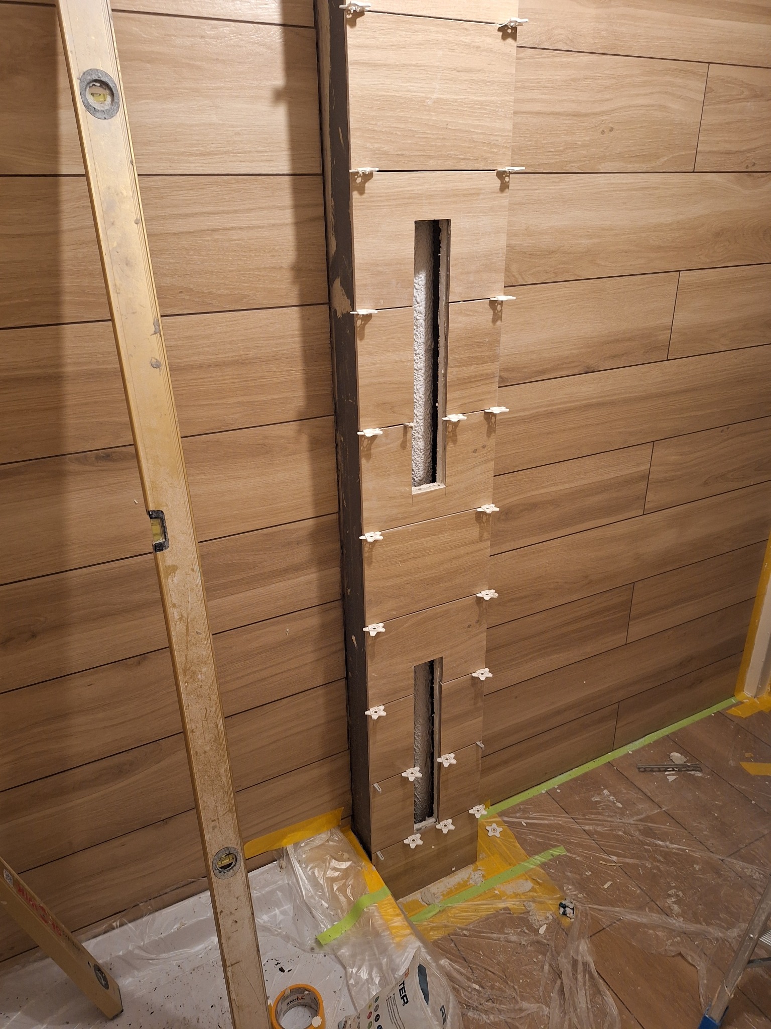 Układanie drewnopodobnych płytek na ścianie z pionowym wnęką, widoczne krzyżyki dystansowe, poziomica oparta o ścianę, podłoga zabezpieczona folią malarską.