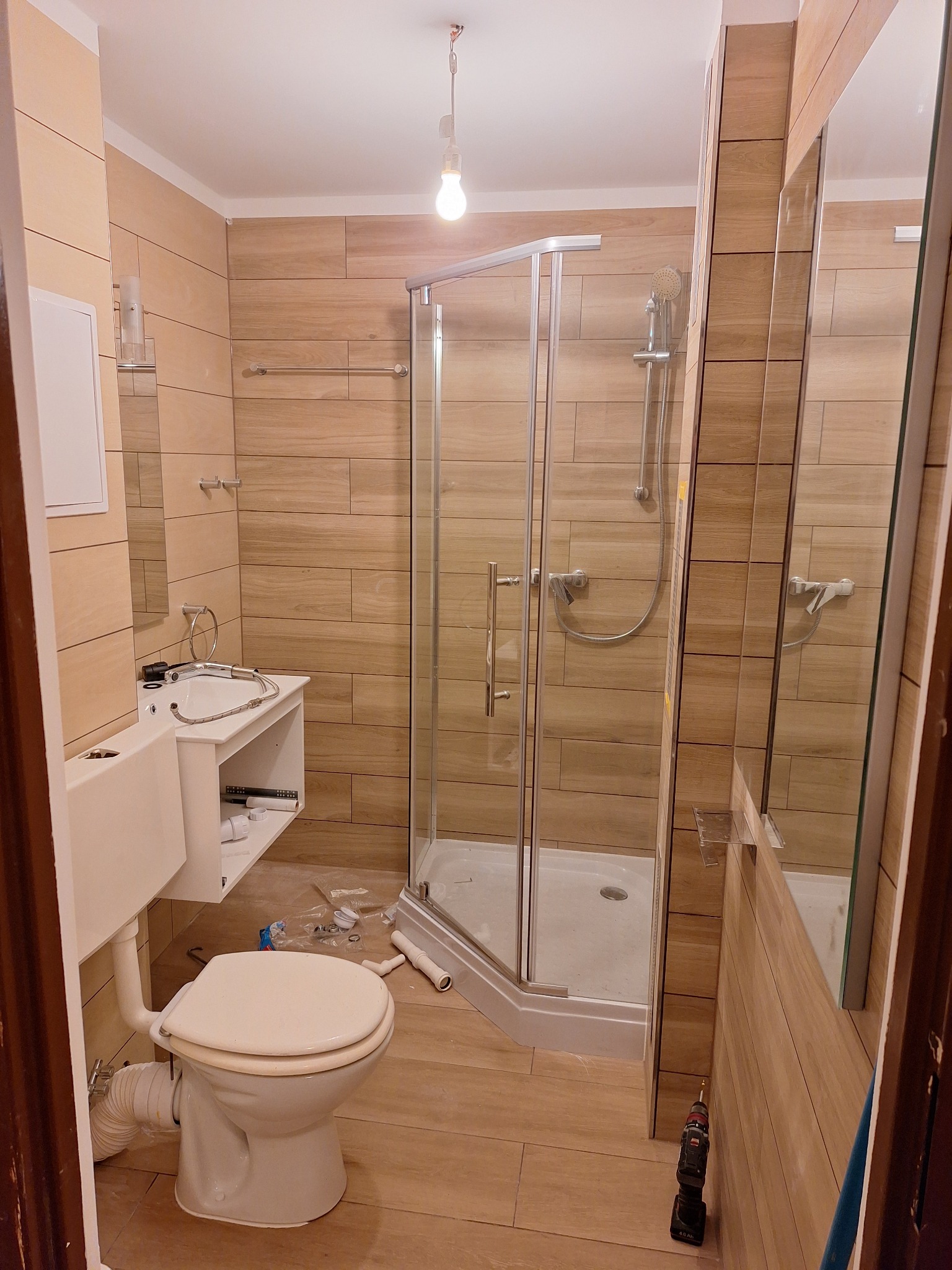 Niedokończona łazienka z kabiną prysznicową, umywalką z szafką i toaletą, płytki imitujące drewno na ścianach, widoczne narzędzia i akcesoria montażowe.