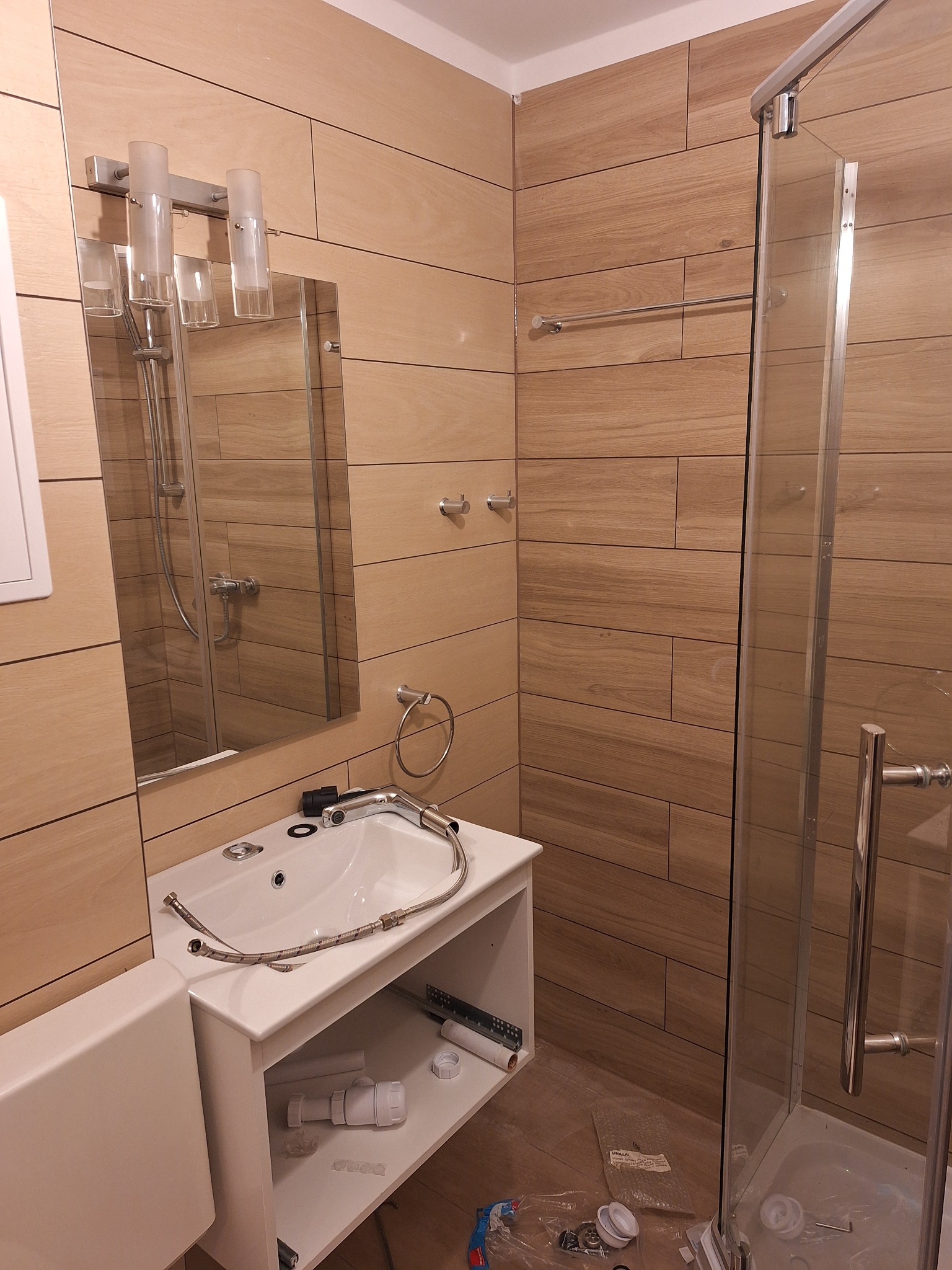 Montaż umywalki w łazience z drewnopodobnymi płytkami na ścianach, widoczna kabina prysznicowa i elementy instalacji hydraulicznej