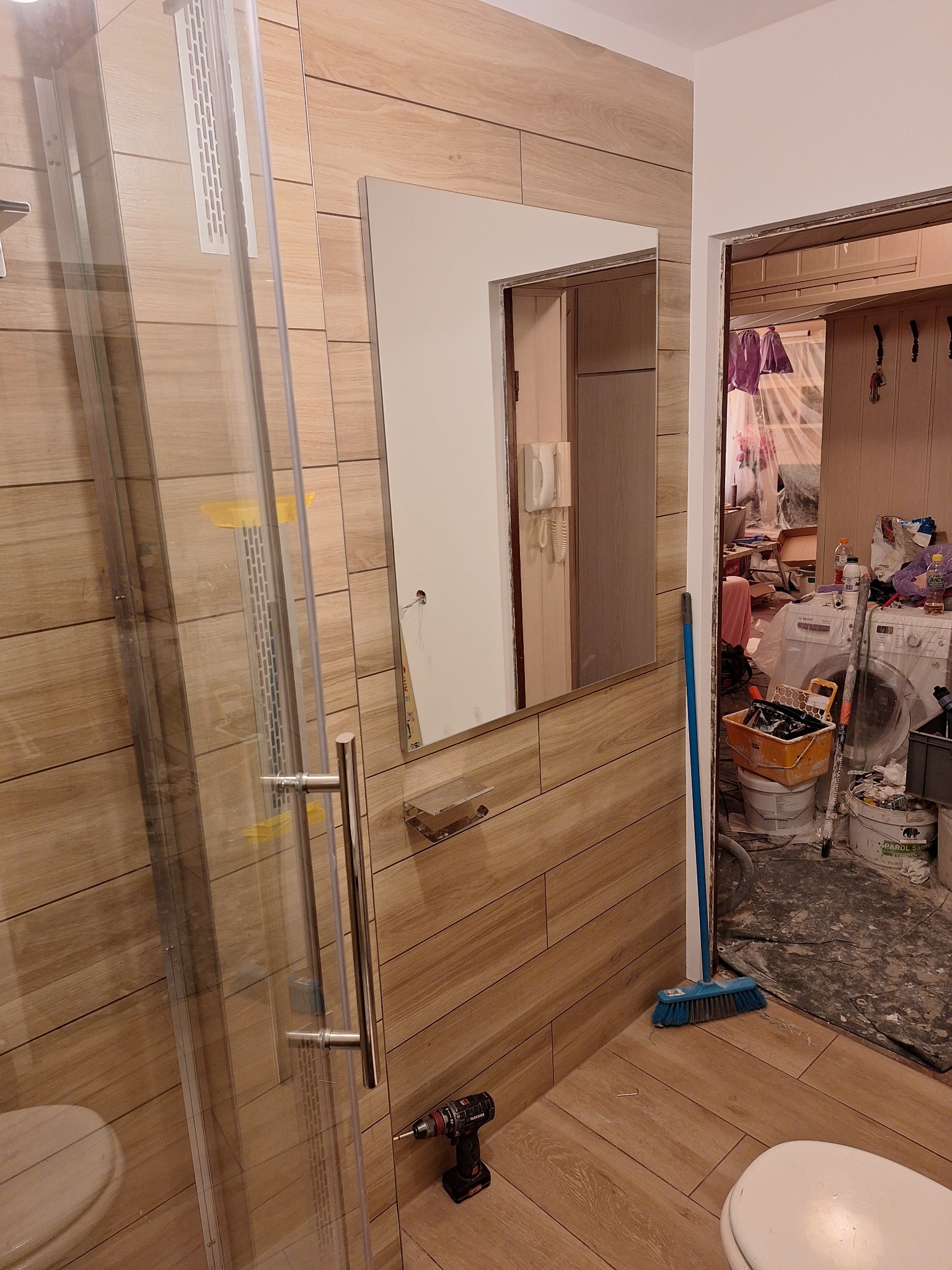 Fragment łazienki w trakcie prac: widoczna kabina prysznicowa z zamontowanym panelem, lustro nad umywalką, płytki imitujące drewno na ścianach, w tle bałagan remontowy i pralka osłonięta folią,...