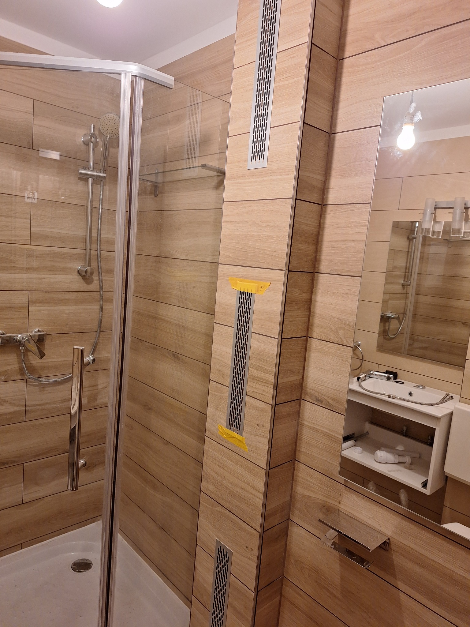 Wykończona łazienka z kabiną prysznicową, umywalką z szafką, lustrem i drewnopodobnymi płytkami na ścianach, widoczne żółte taśmy zabezpieczające na kratkach wentylacyjnych.