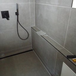 GawBud - Nowoczesna kabina prysznicowa z szarymi płytkami, czarną armaturą i ukrytą rewizją, widoczne folie ochronne na półce.