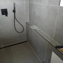 Nowoczesna kabina prysznicowa z szarymi płytkami, czarną armaturą i ukrytą rewizją, widoczne folie ochronne na półce.