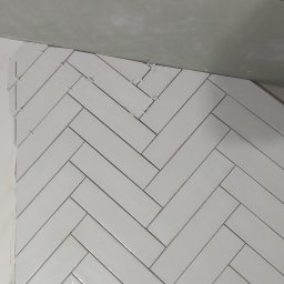 GawBud - Białe, błyszczące płytki ułożone w jodełkę na ścianie, z widocznymi krzyżykami dystansowymi, w trakcie prac wykończeniowych.