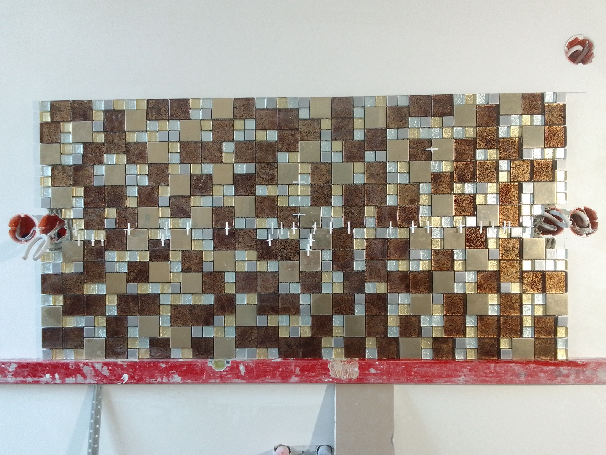 Mozaika z brązowych, beżowych i srebrnych płytek kwadratowych na ścianie, w trakcie montażu, z krzyżykami dystansowymi i poziomicą w kolorze czerwonym.