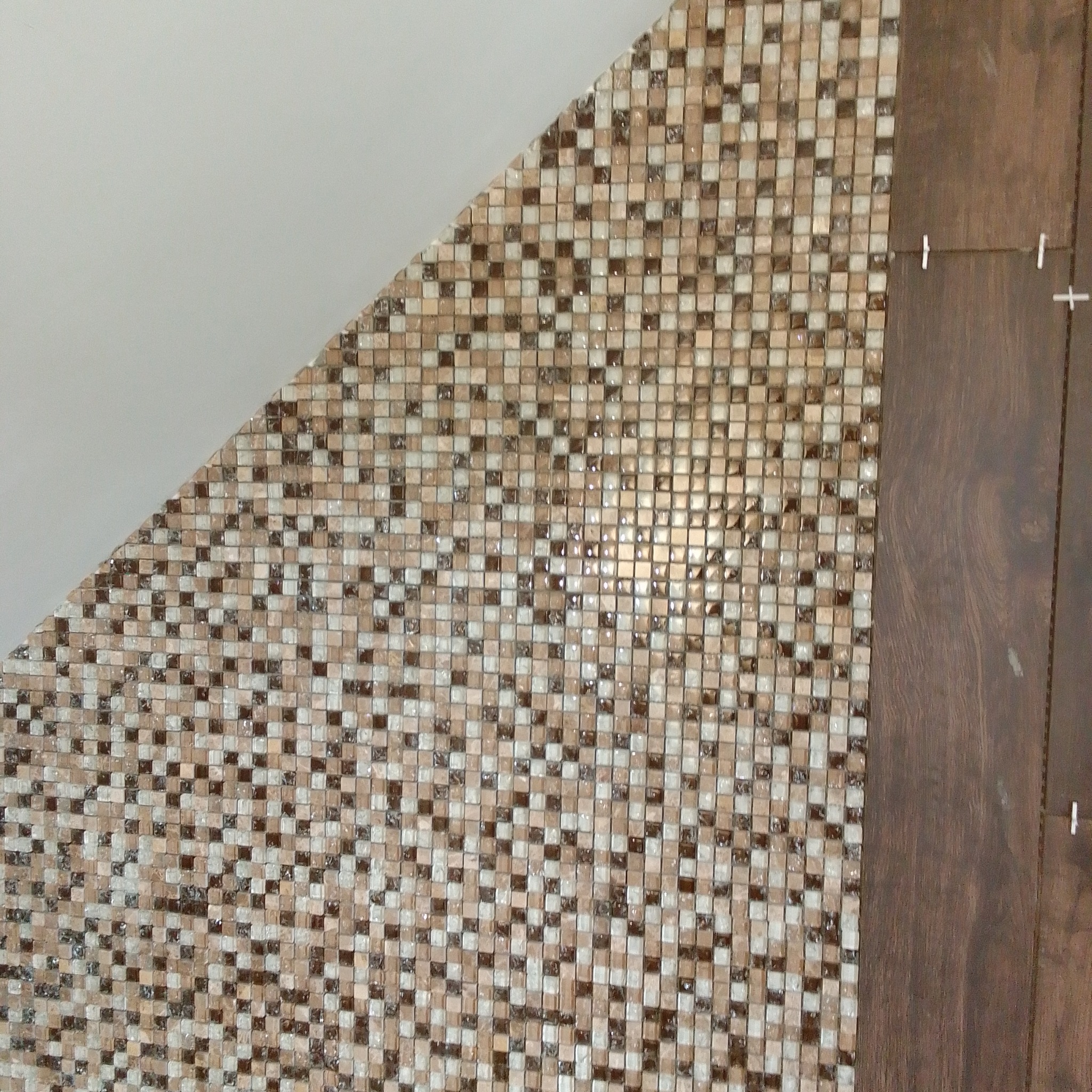 Układanie mozaiki w odcieniach brązu i beżu przy krawędzi z ciemnym drewnem, widoczne krzyżyki dystansowe.