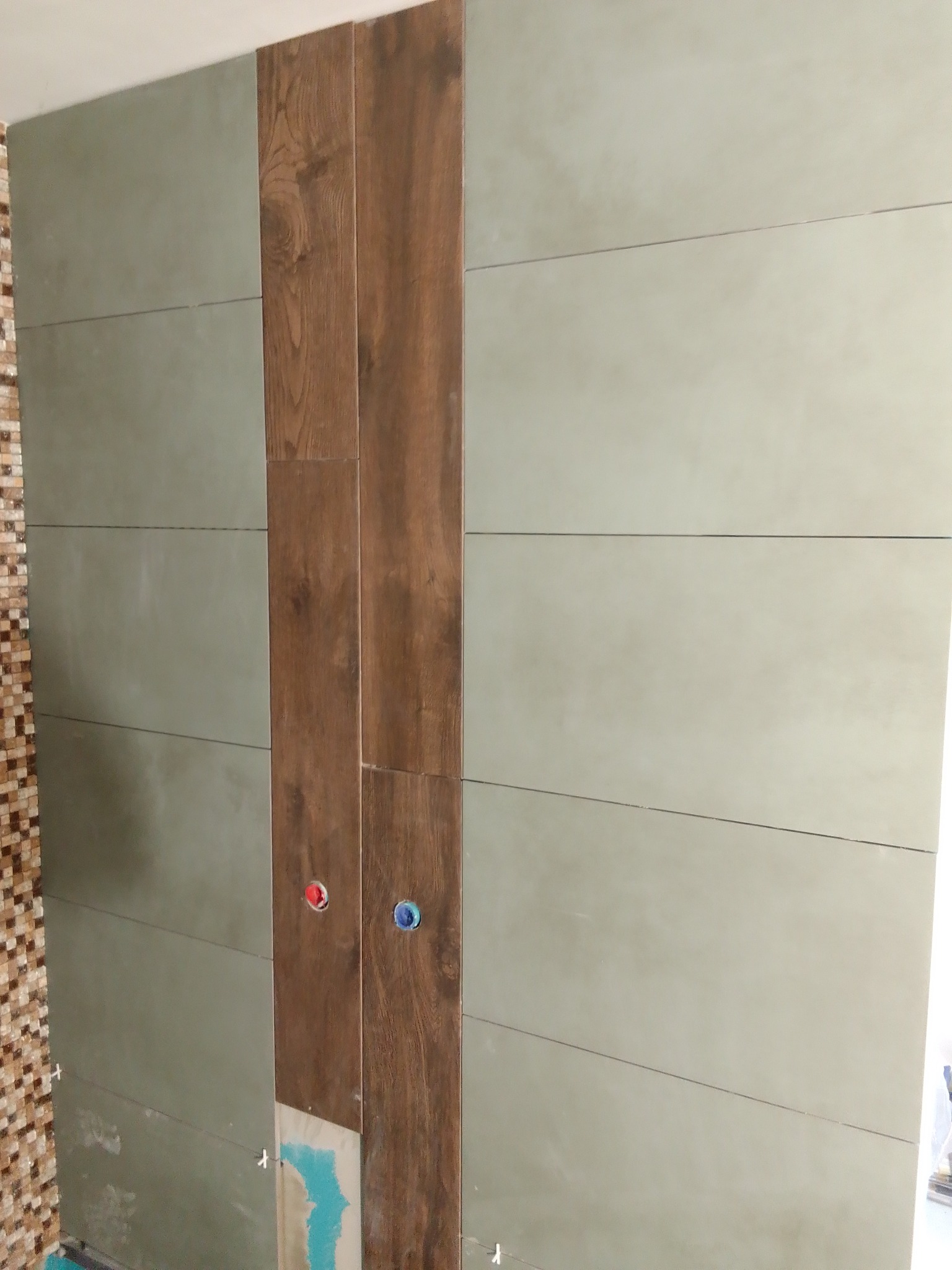 Ściana w trakcie wykańczania z poziomymi, szarymi płytkami i pionowym pasem płytek imitujących drewno, widoczne otwory na rury i krzyżyki dystansowe.