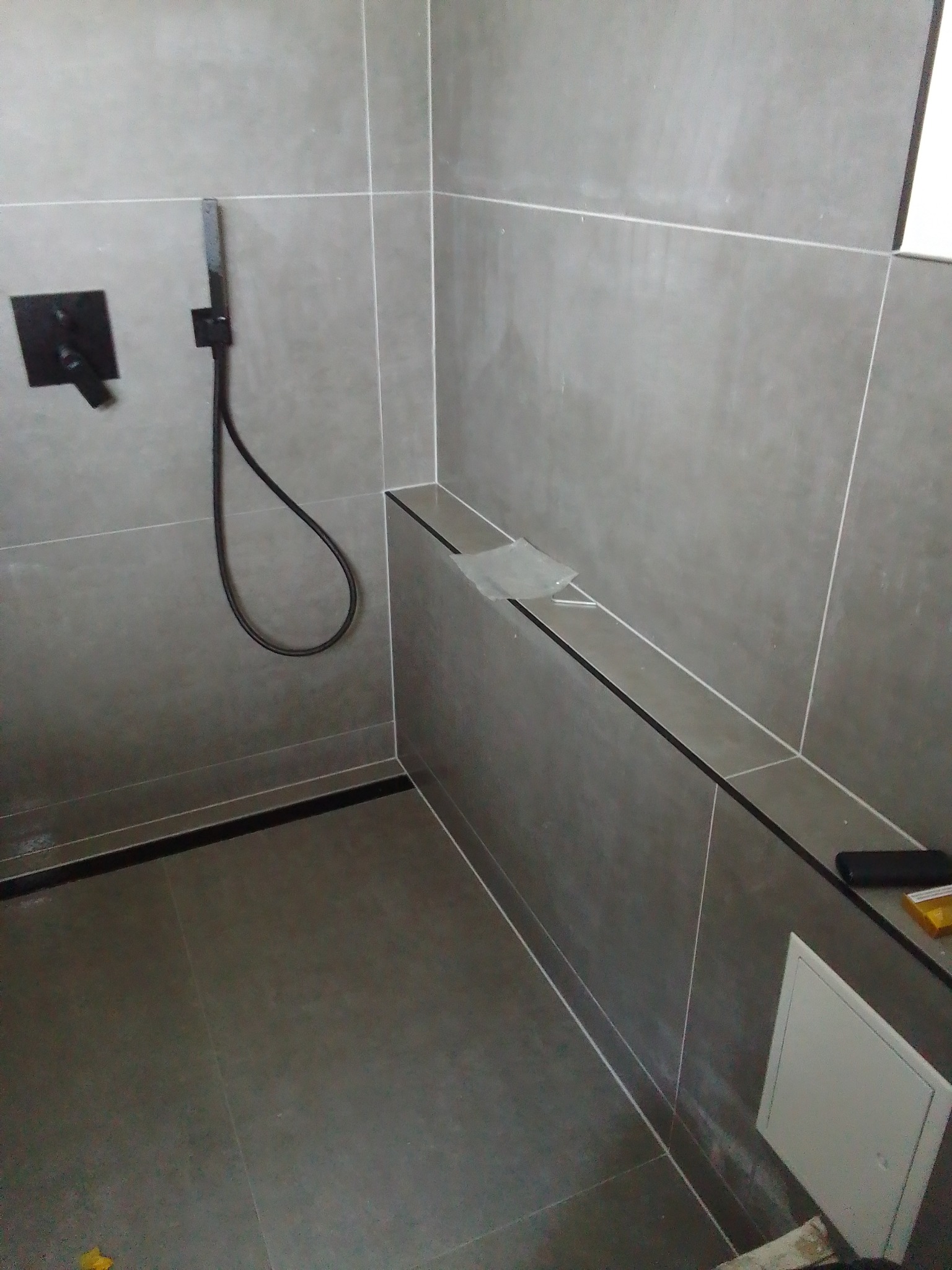 Nowoczesna kabina prysznicowa z szarymi płytkami, czarną armaturą i ukrytą rewizją, widoczne folie ochronne na półce.