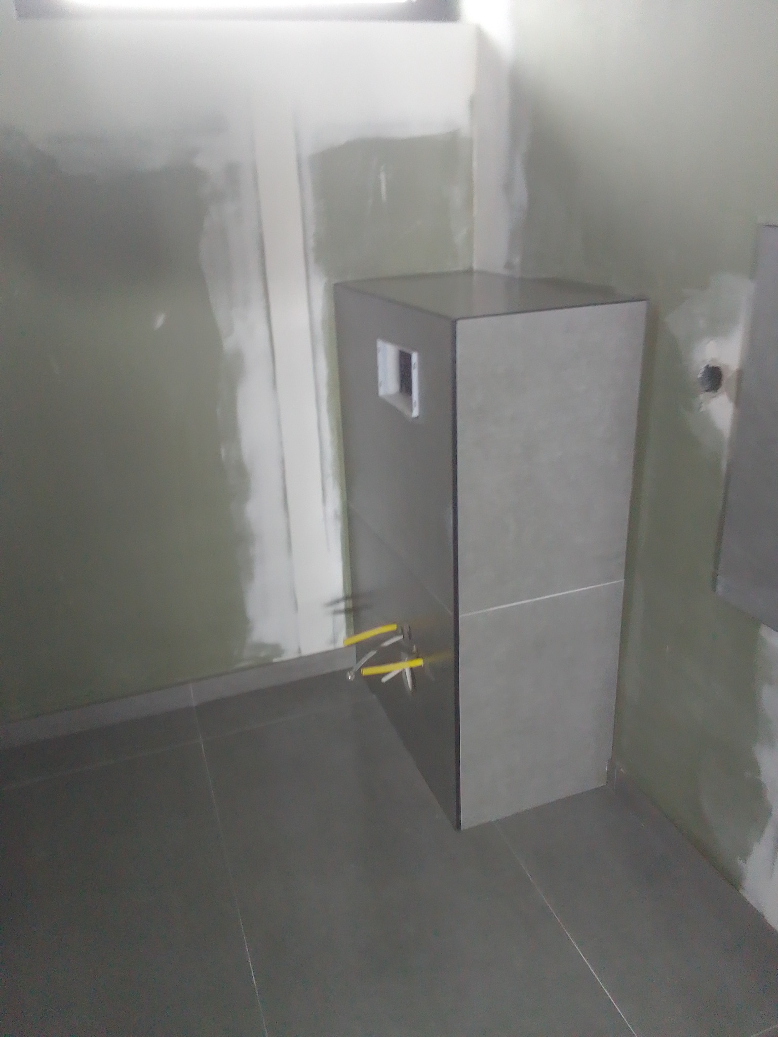 Stelaż podtynkowy WC obudowany płytkami w szarej tonacji, widoczne wystające rury instalacyjne, ściany częściowo wykończone gładzią, ciemna podłoga z płytek.