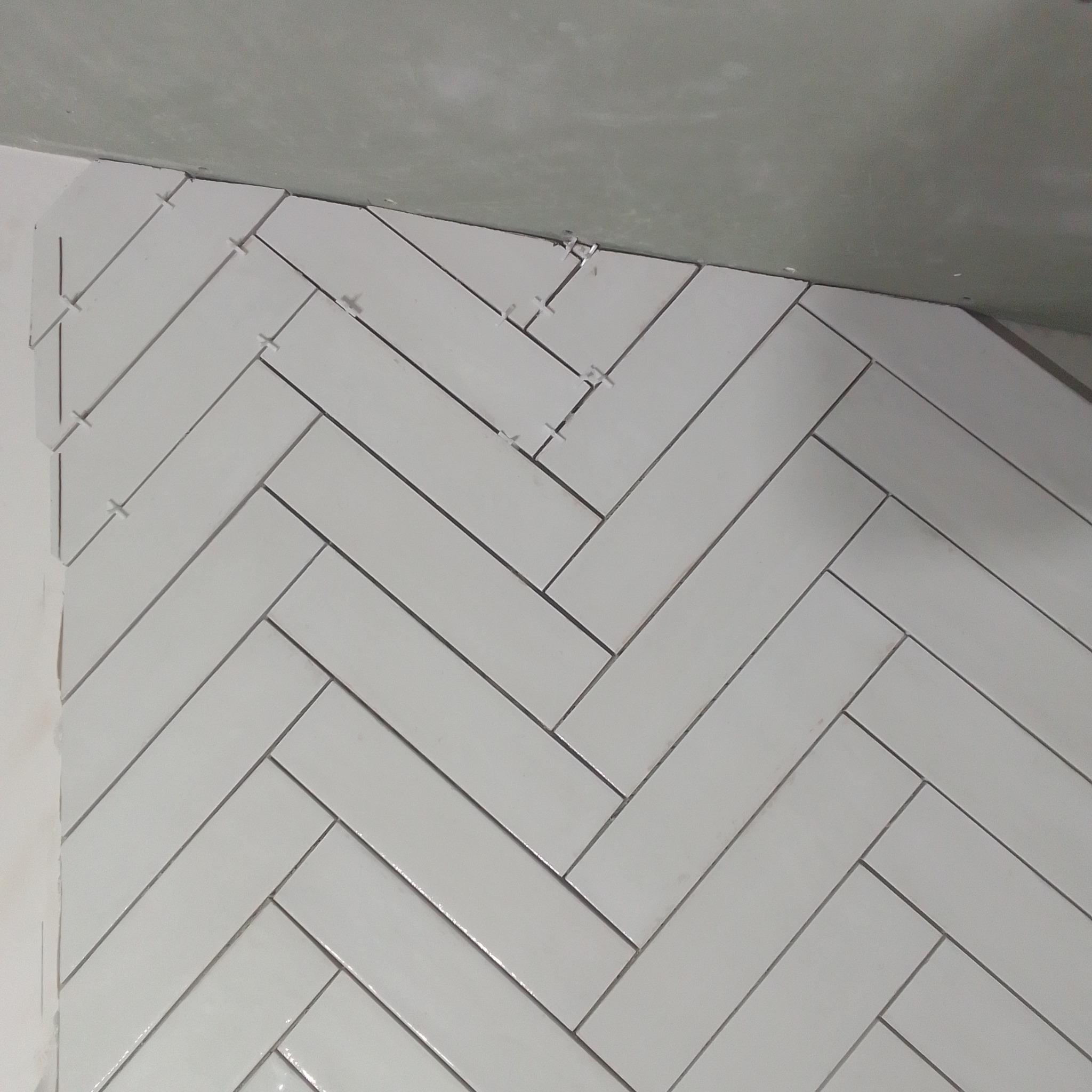 Białe, błyszczące płytki ułożone w jodełkę na ścianie, z widocznymi krzyżykami dystansowymi, w trakcie prac wykończeniowych.