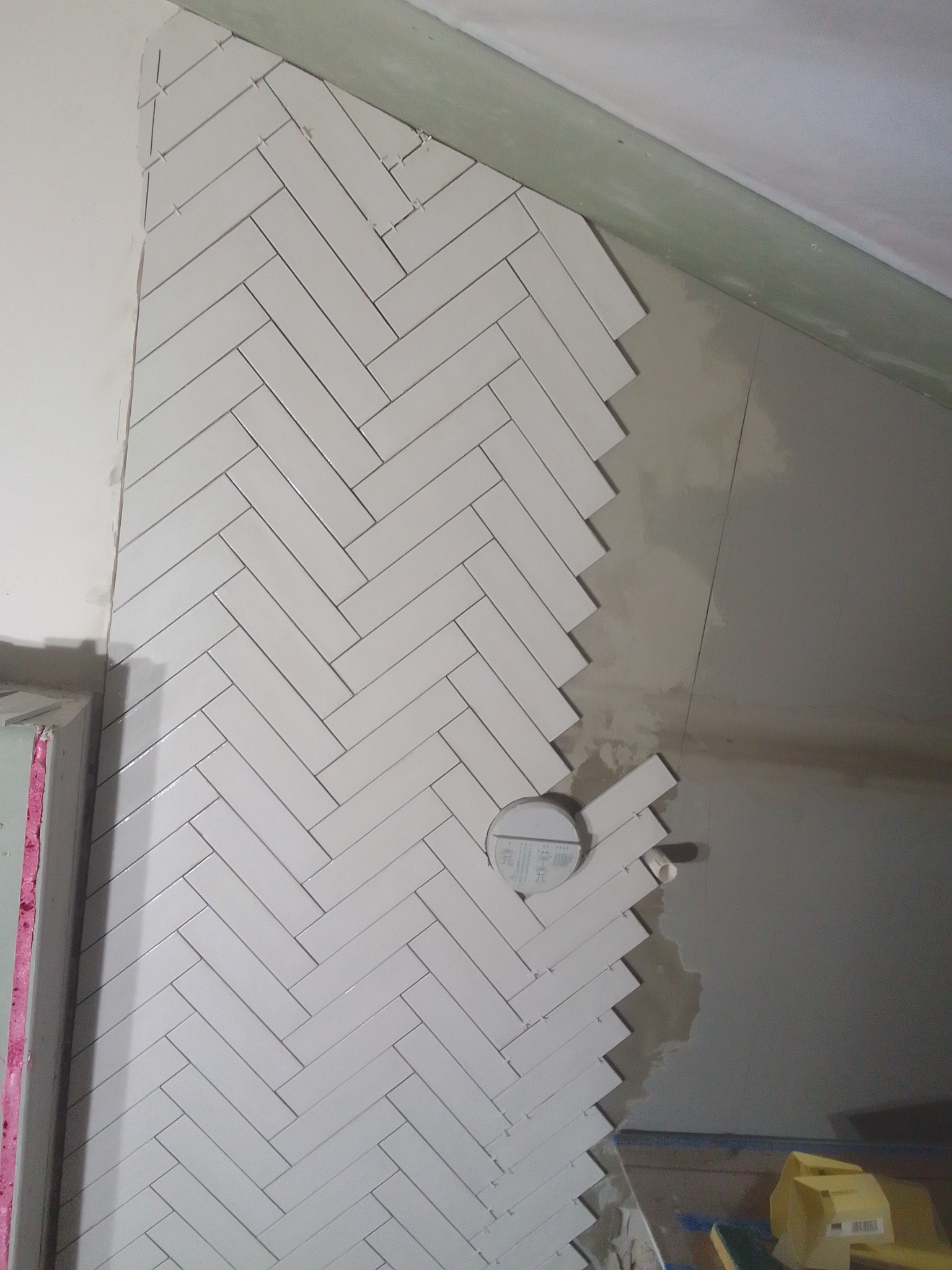 Ściana w trakcie układania białych, podłużnych płytek w jodełkę, widoczne krzyżyki dystansowe i niedokończone krawędzie przy suficie i ścianie.