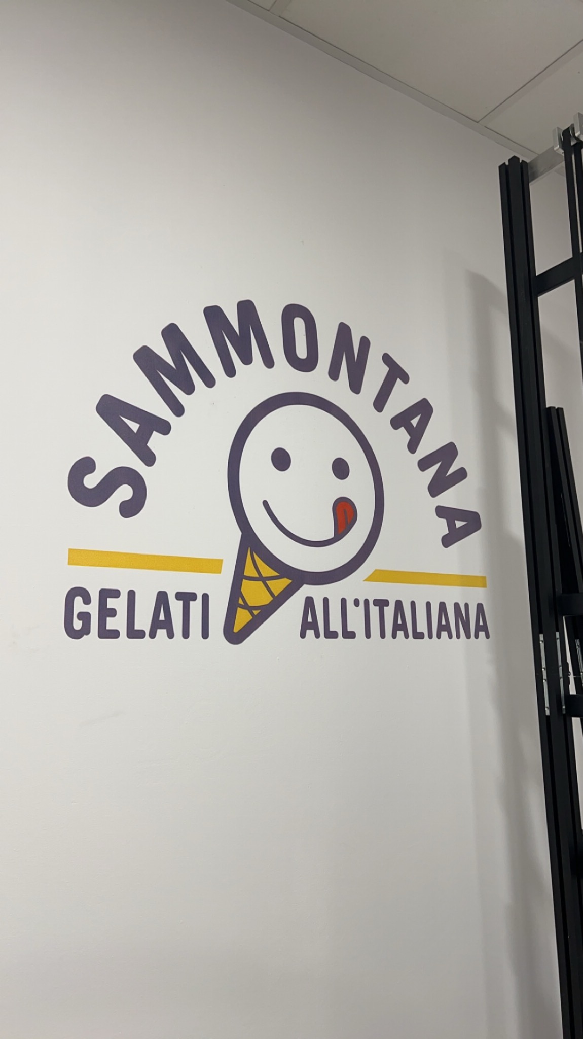 Świeżo wytapetowana ściana z naklejonym logo lodziarni Sammontana Gelati All'Italiana, przedstawiającym uśmiechniętą buźkę w rożku oblizującą się na białym tle, z czarnymi elementami rusztowania po...