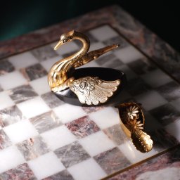 Wiktoria Żółciak - Dekoracyjna, pozłacana figurka łabędzia z czarną podstawą i otwieranym pojemnikiem w kształcie łódki, stojąca na marmurowej szachownicy.