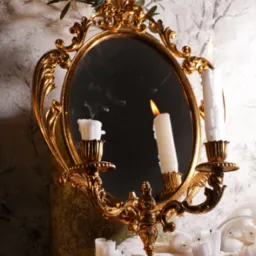 Dekoracyjne, złote lustro owalne z palącymi się świecami w świecznikach, odbijające płomień i dym, na tle marmurkowej ściany i gałązek oliwnych, sfotografowane w Krakowie.