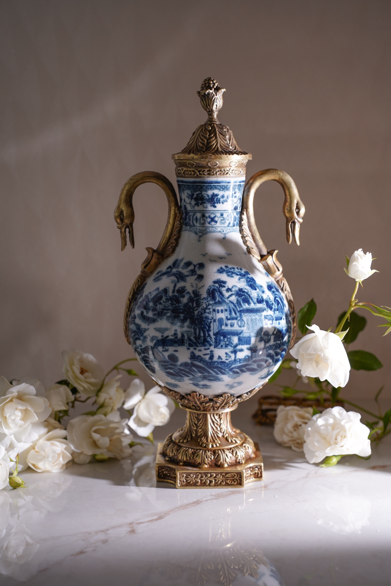Dekoracyjny wazon z białej porcelany z niebieskim wzorem krajobrazowym, zdobiony pozłacanymi elementami w kształcie łabędzi i kwiatów, otoczony białymi różami na marmurowym blacie.