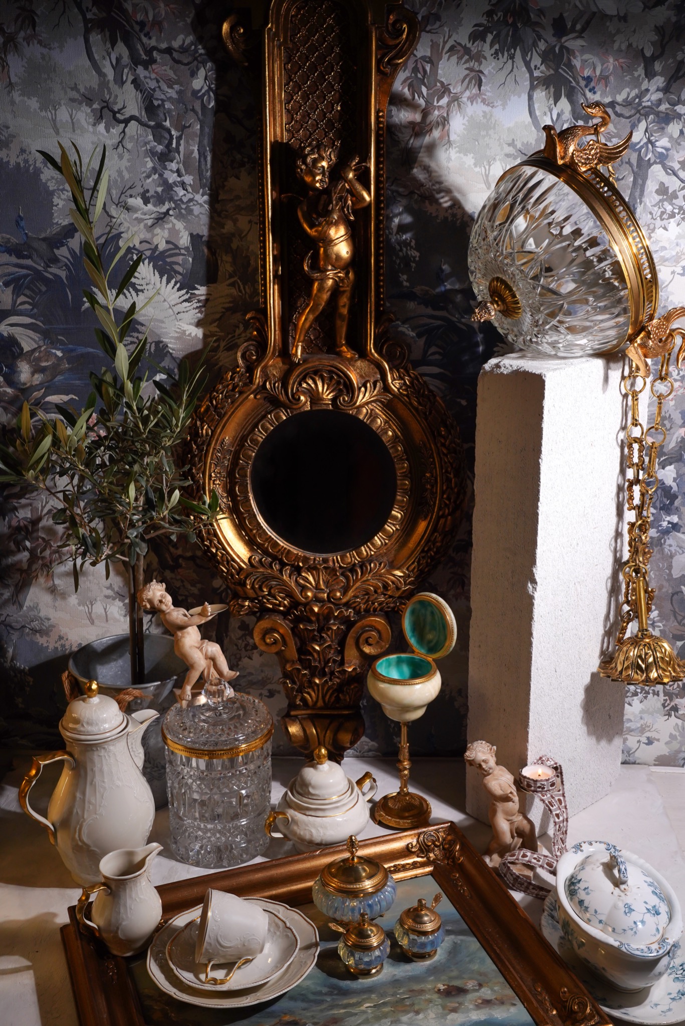 Aranżacja stołu z antycznymi naczyniami, złotym lustrem z figurką anioła, kryształową kulą i rośliną na tle tapety w leśny motyw.