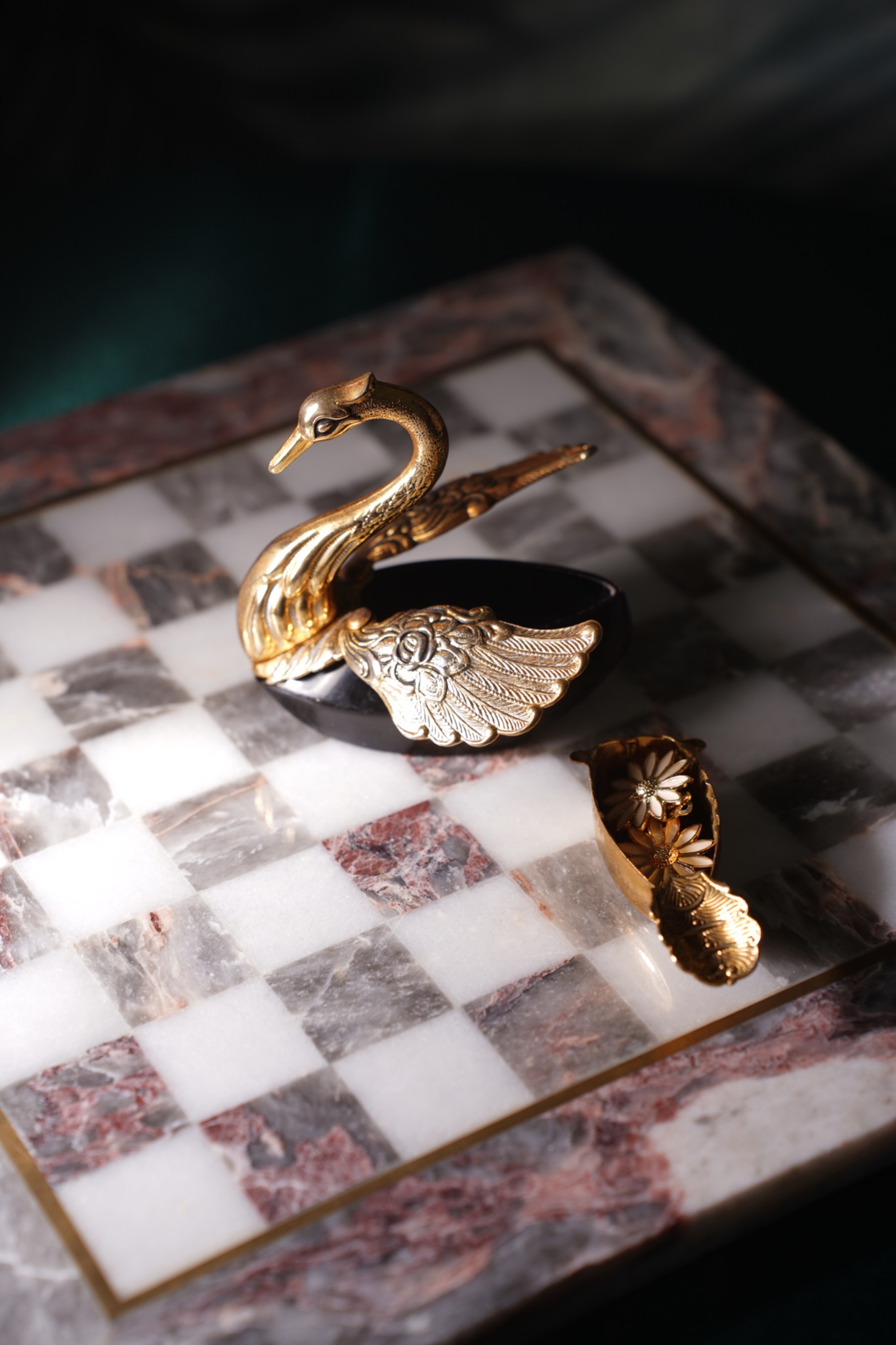 Dekoracyjna, pozłacana figurka łabędzia z czarną podstawą i otwieranym pojemnikiem w kształcie łódki, stojąca na marmurowej szachownicy.