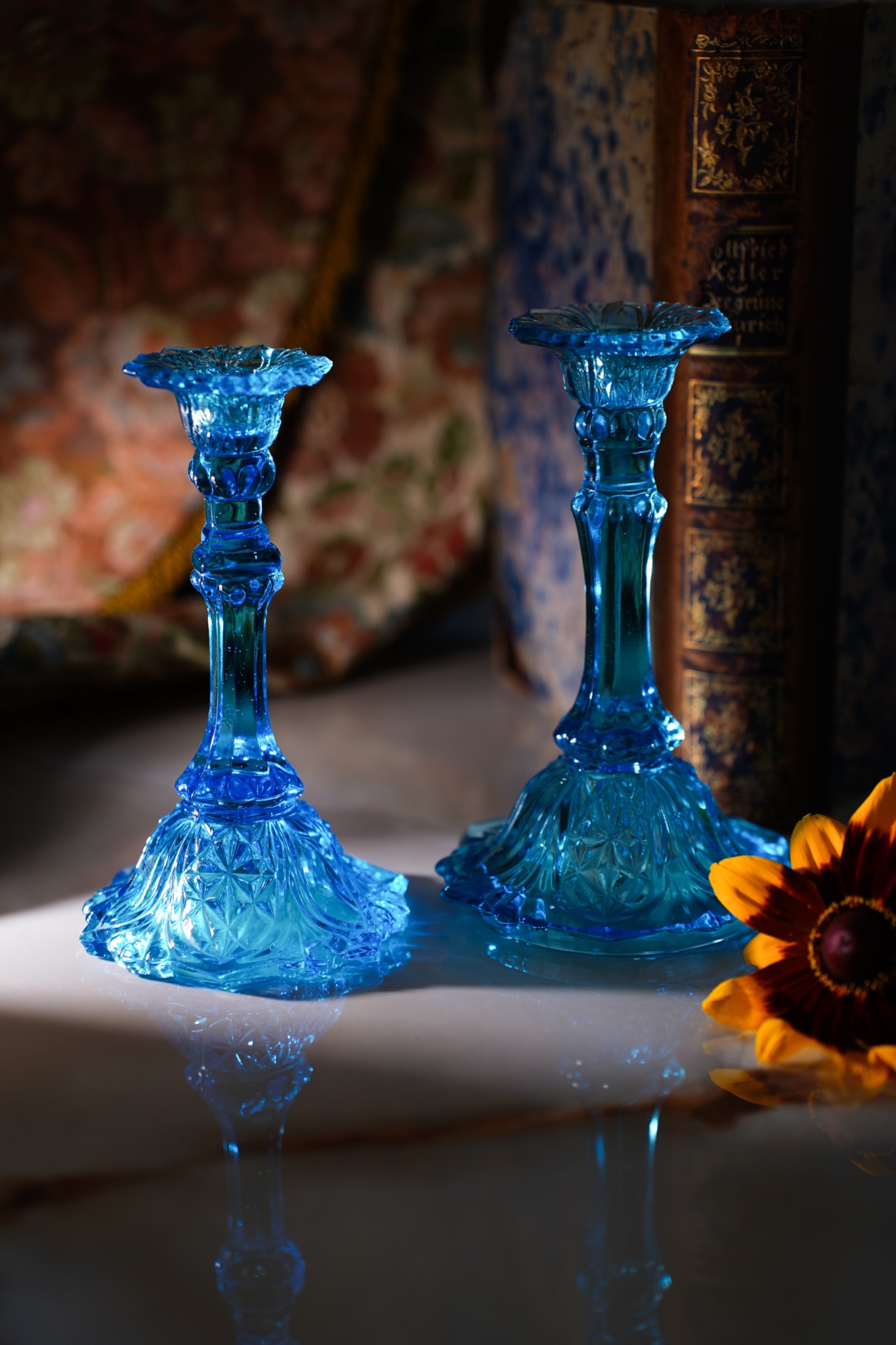 Dwa błękitne, szklane świeczniki na nóżce, z kwiatem rudbekii i starymi książkami w tle, oświetlone ciepłym światłem.