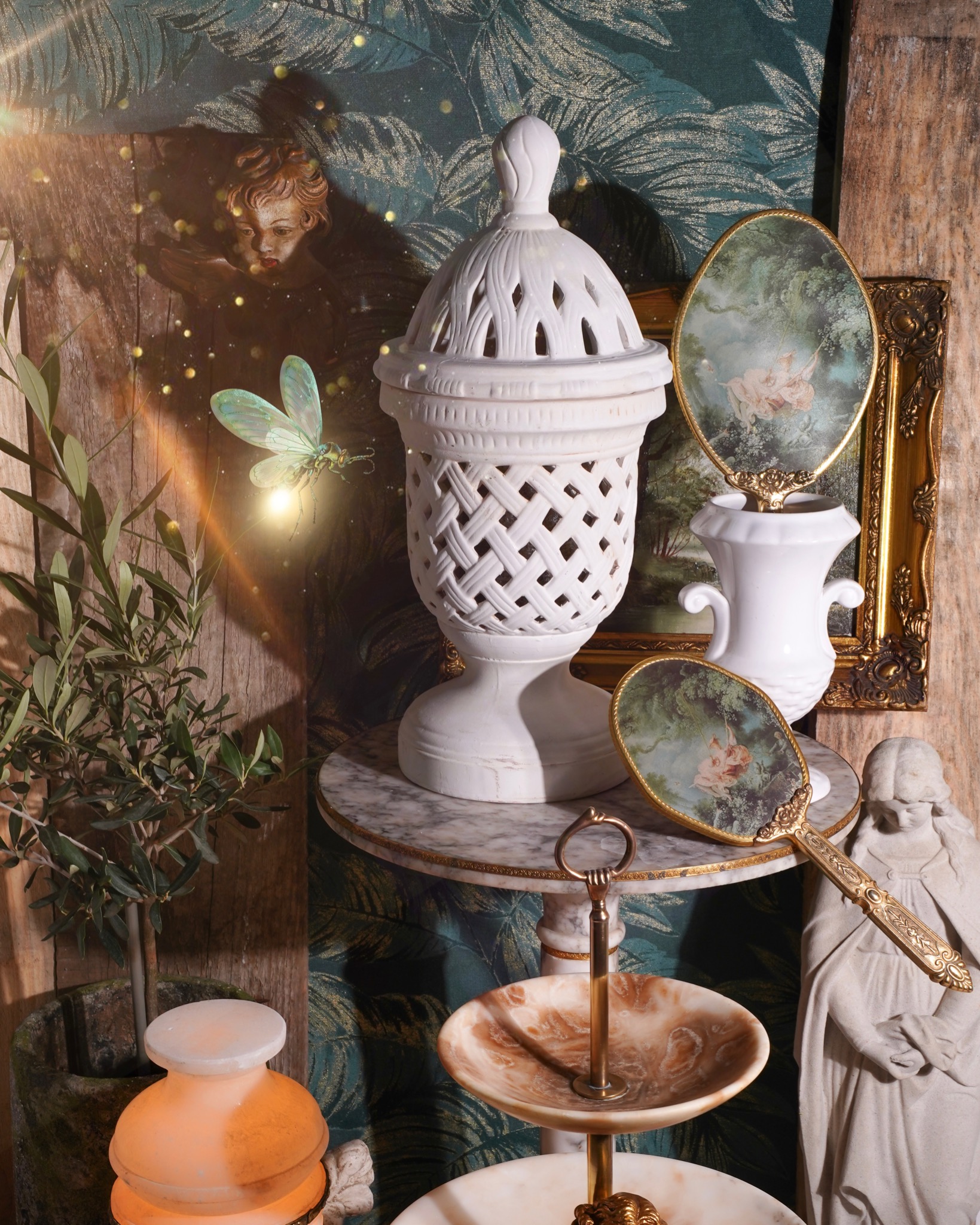 Aranżacja wnętrza z dekoracjami w stylu vintage: biała ażurowa lampion, marmurowy stolik, lustra z motywem huśtawki, figurka anioła, roślina w doniczce i świetlik z aureolą.