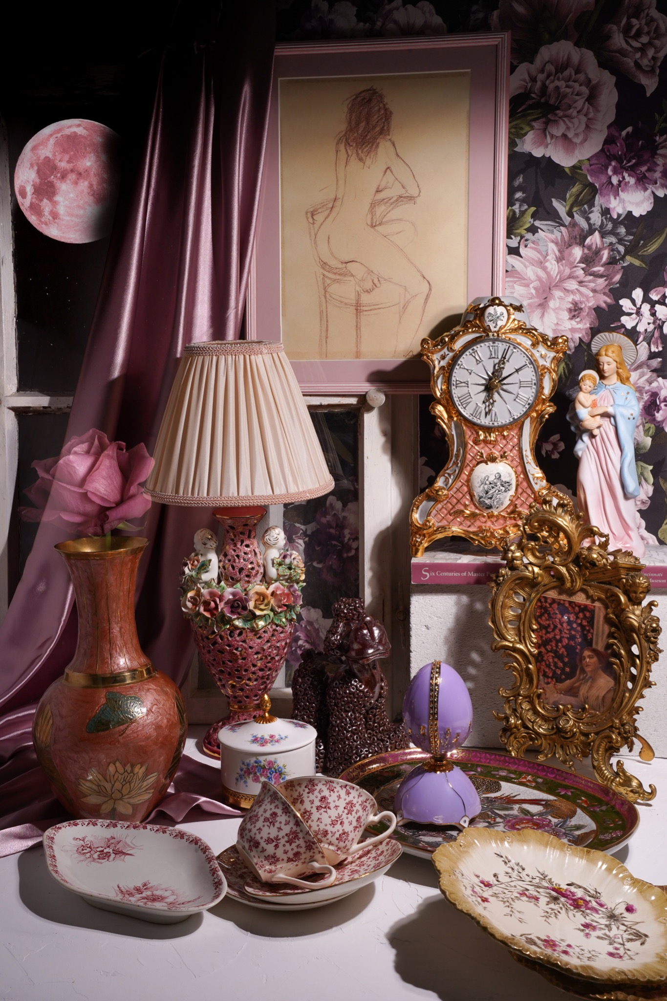 Martwa natura z antykami: wazon z różą, lampa z aniołkami, zegar kominkowy, figurka Madonny z Dzieciątkiem, filiżanki w róże, talerze dekoracyjne, rysunek kobiety i księżyc na ścianie.