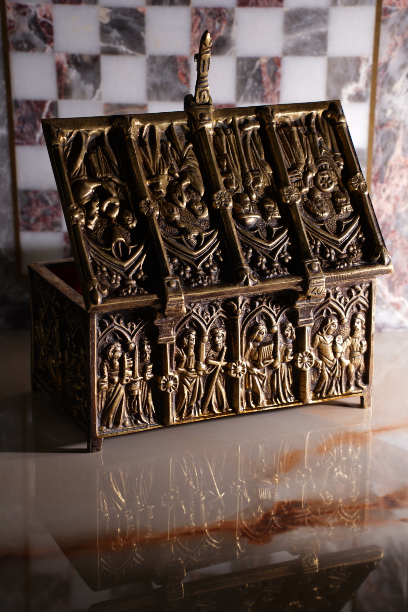 Otwarta, zdobiona, metalowa szkatułka z reliefami postaci na marmurowym blacie, odbijająca się w powierzchni.