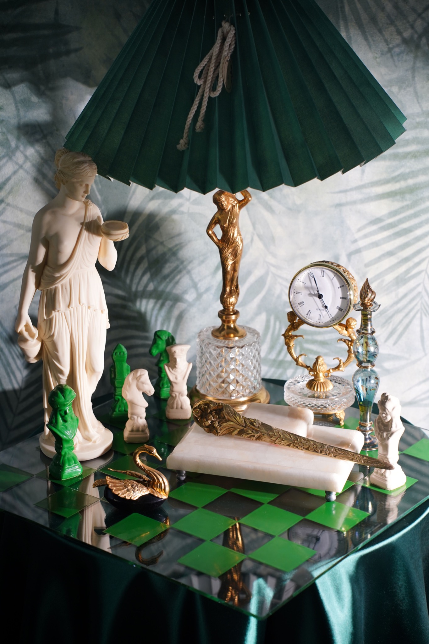 Martwa natura z antycznymi figurkami, zegarem i dekoracyjnymi przedmiotami w odcieniach złota, bieli i zieleni, ustawionymi na szachownicy pod zielonym abażurem lampy.