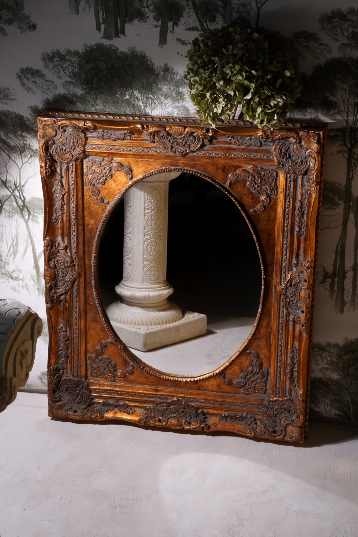 Złota, dekoracyjna rama z owalnym otworem, ustawiona na podłodze, z kolumną widoczną wewnątrz otworu, na tle ściany z motywem drzew i bukietem hortensji.