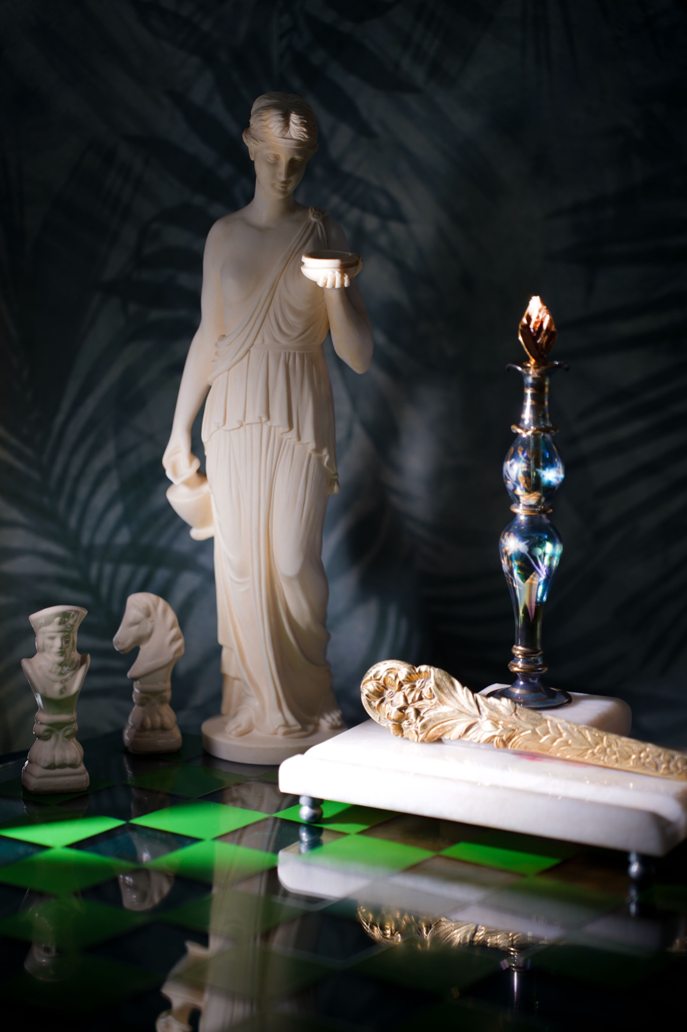 Martwa natura z białą statuetką kobiety trzymającej naczynie, szachowymi figurkami i ozdobnym, złotym nożem do listów na szachownicy z zielono-czarnymi polami i tle z motywem liści palmowych.