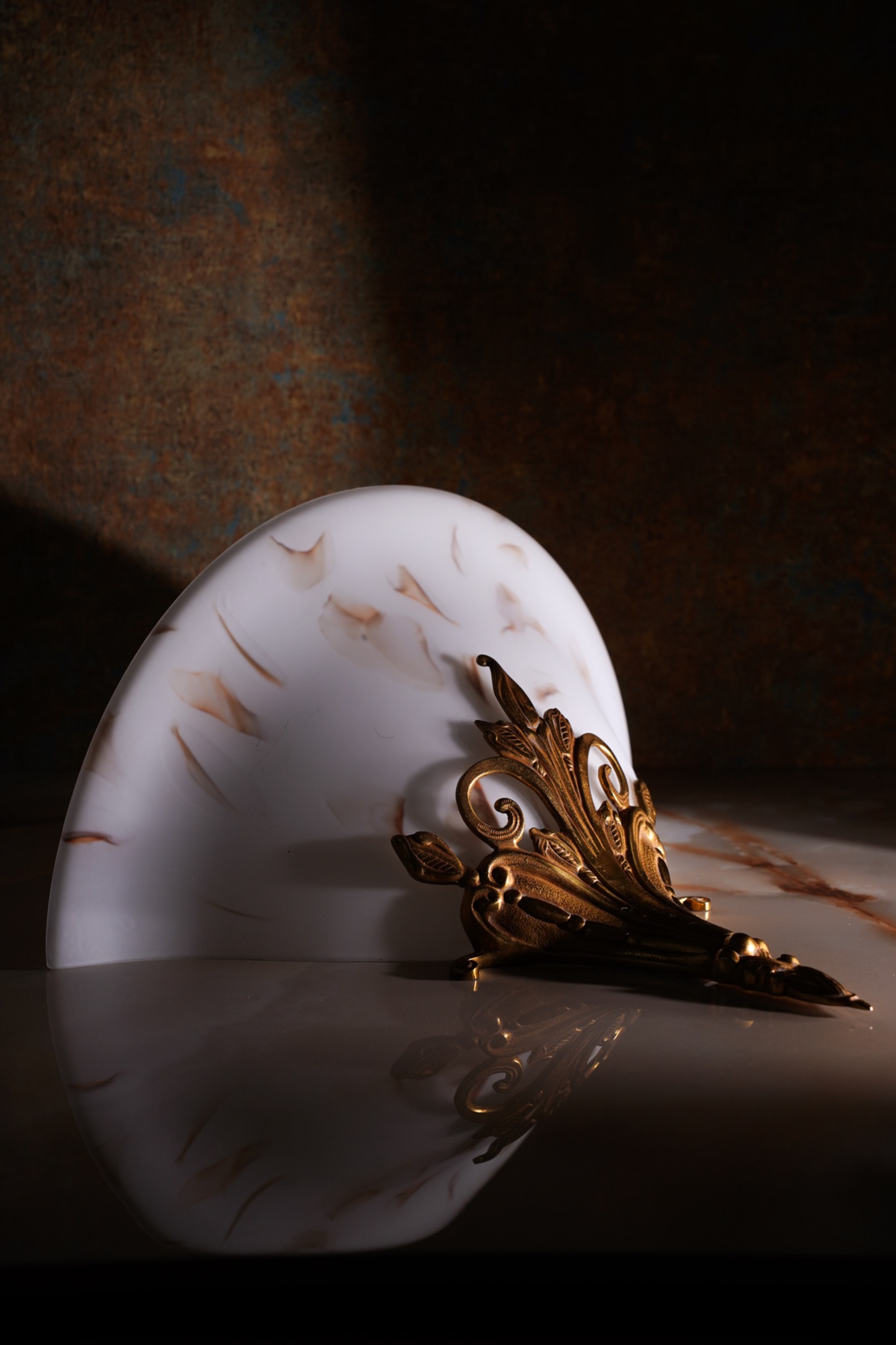 Stylizowana lampa ścienna z białym kloszem z brązowymi smugami i ozdobnym, złotym mocowaniem z liśćmi na marmurowym blacie z ciemnym tłem.